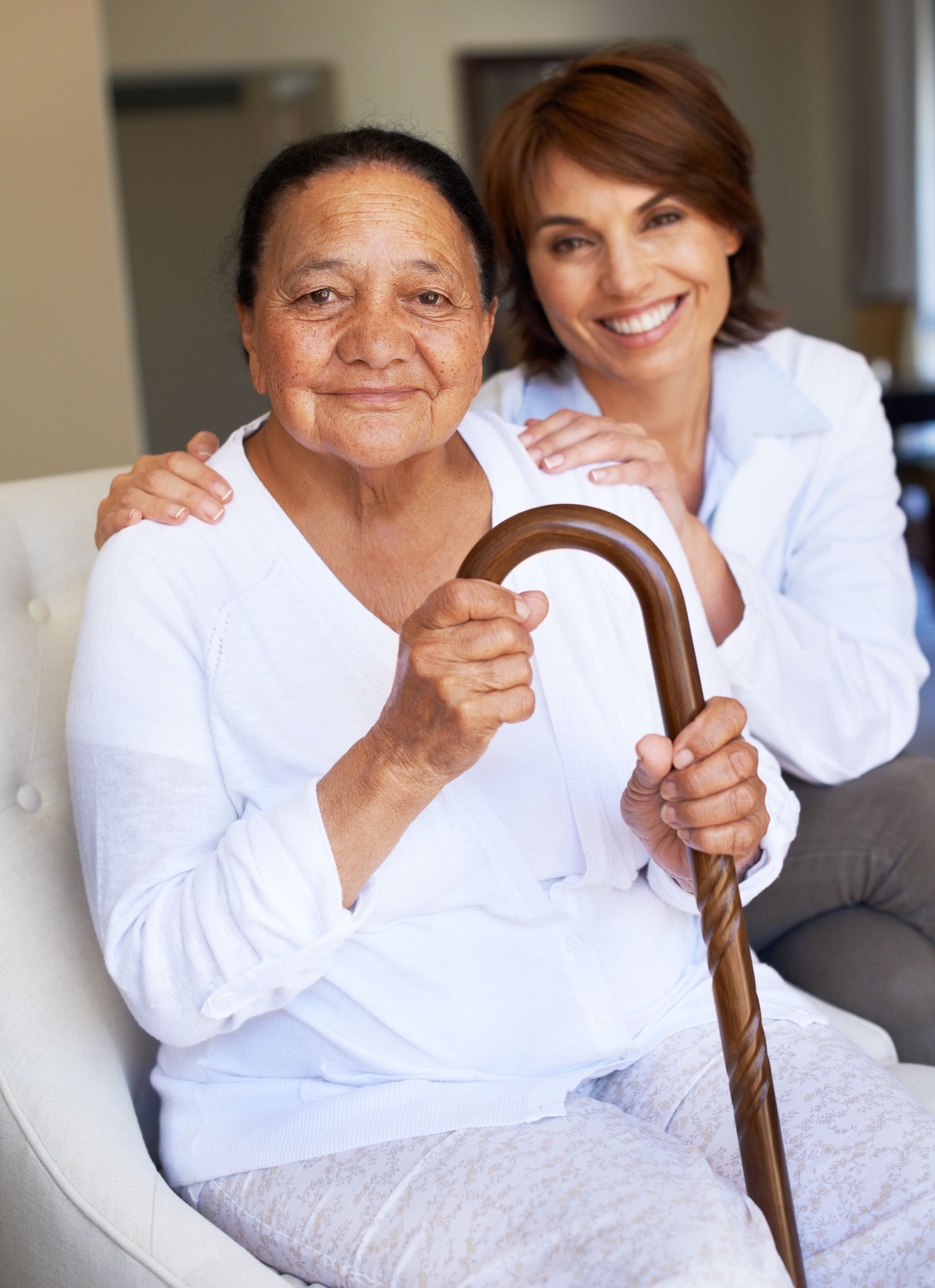 A smiling caregiver with hands on the shoulders of an older person holding a wooden cane, seated in an indoor setting.