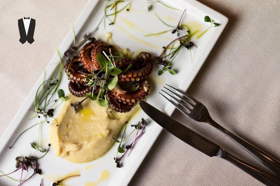 Un piatto rettangolare bianco con tentacoli di polpo grigliati serviti su una purea liscia e guarnito con microverdure.