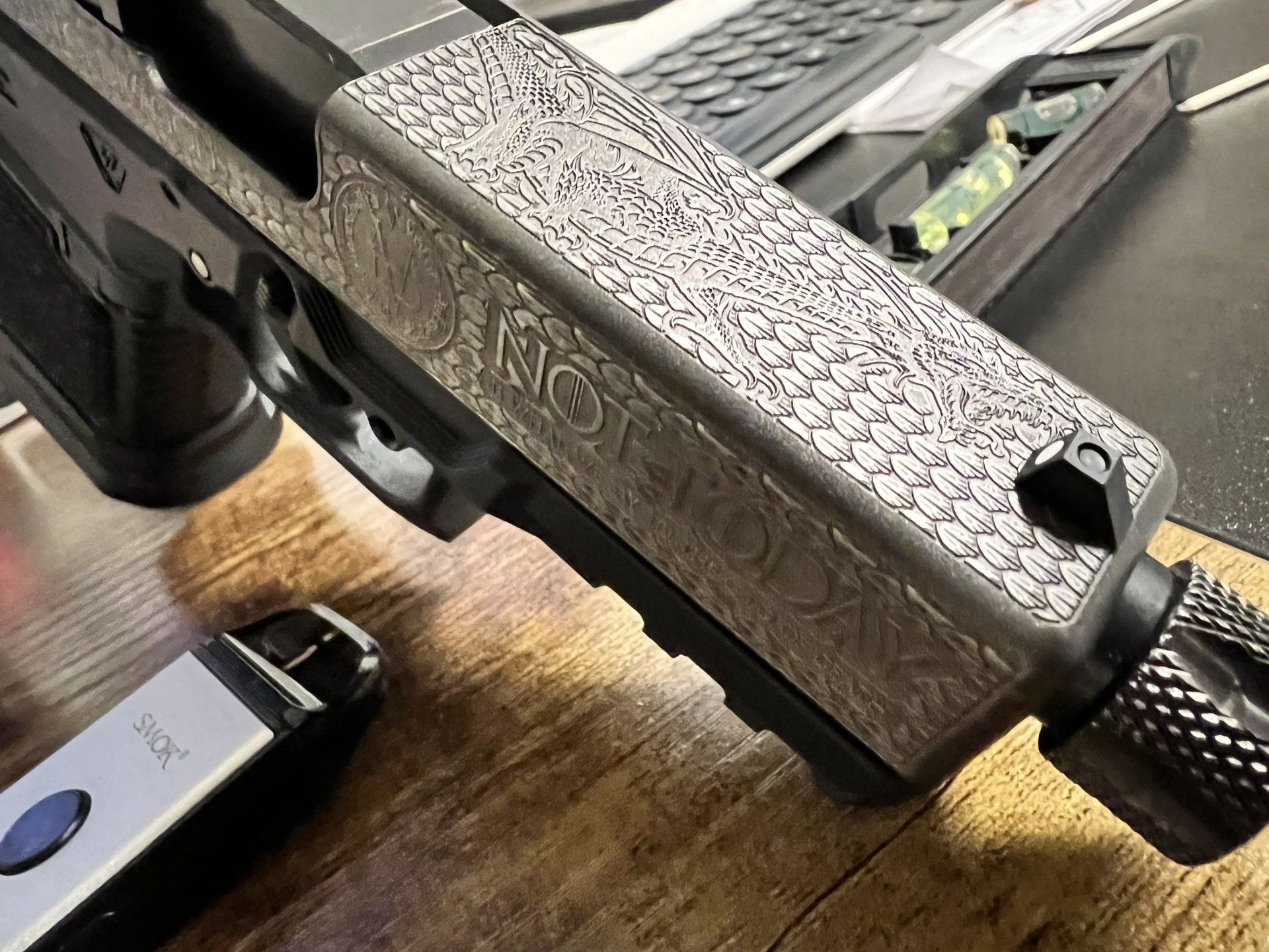 Close-up of a speckled metal firearm slide and textured grip on a wooden table, with a phone nearby