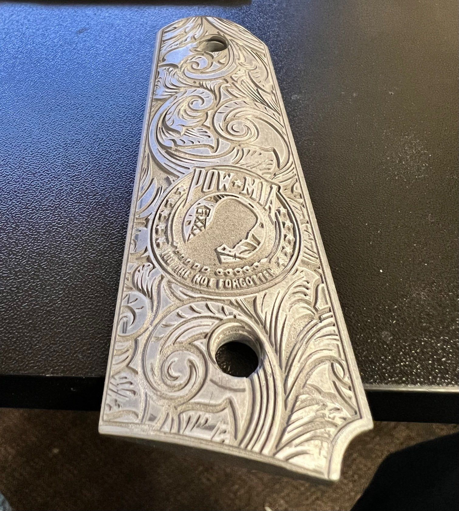 Decorative engraved metal plate with ornate floral scrollwork and two holes on a dark tabletop