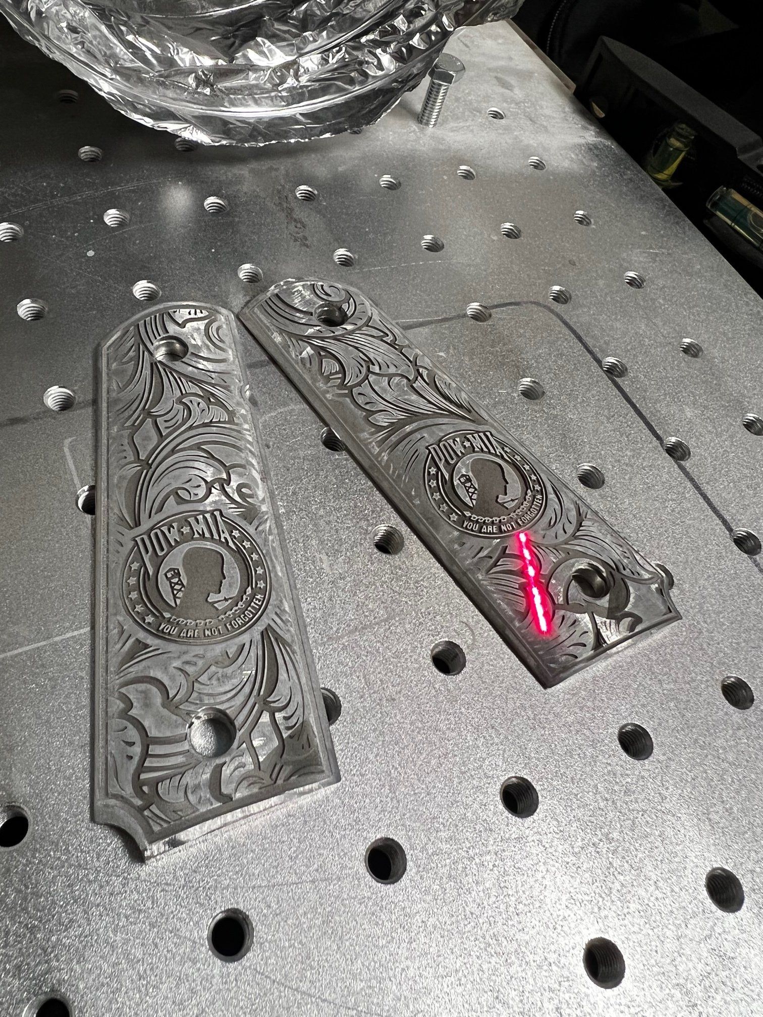 Two engraved metal knife scales on a perforated workbench, one dark and one with a red accent