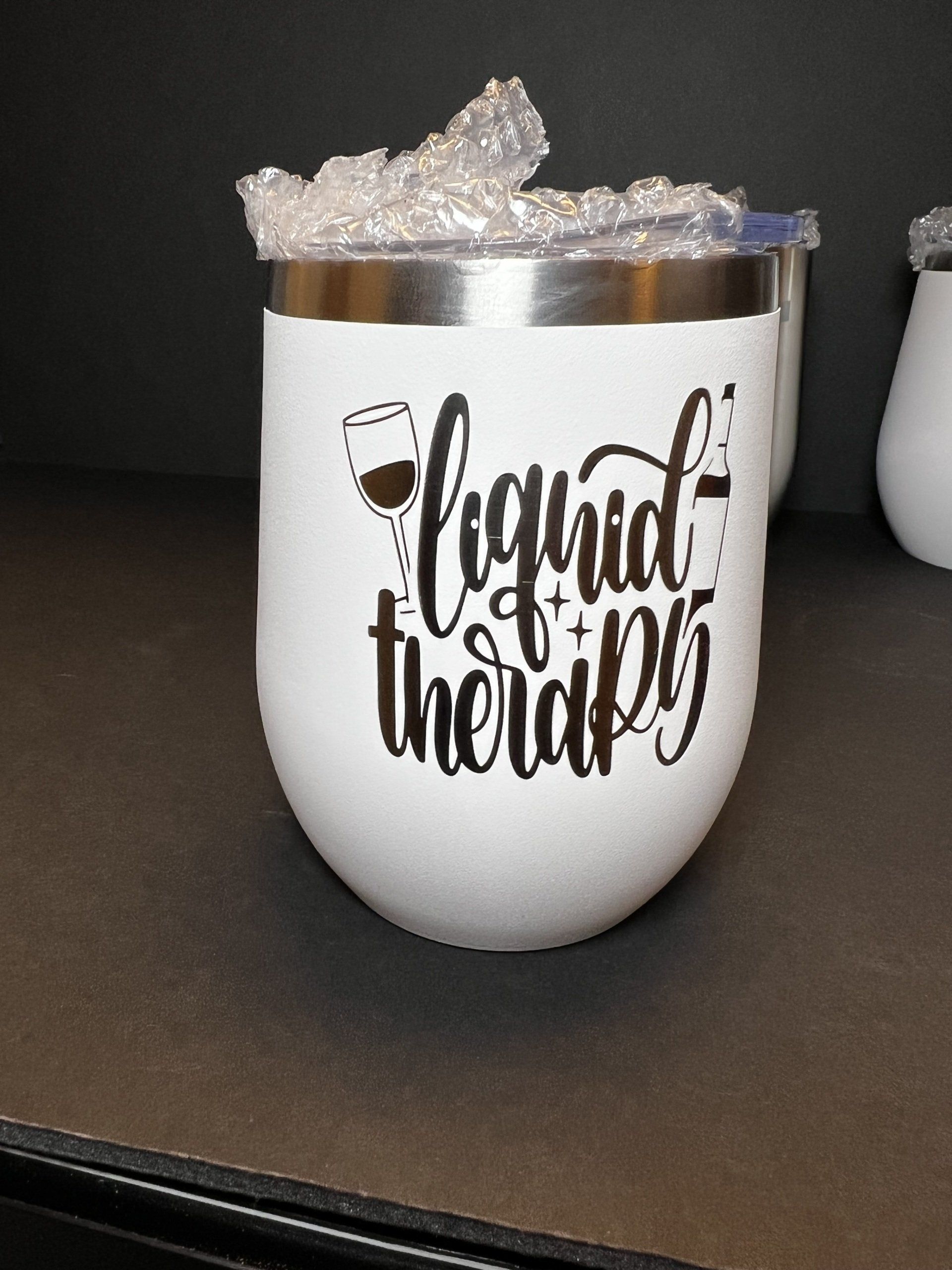 White tumbler with gold rim and “Liquid therapy” text, filled with ice on a dark countertop.