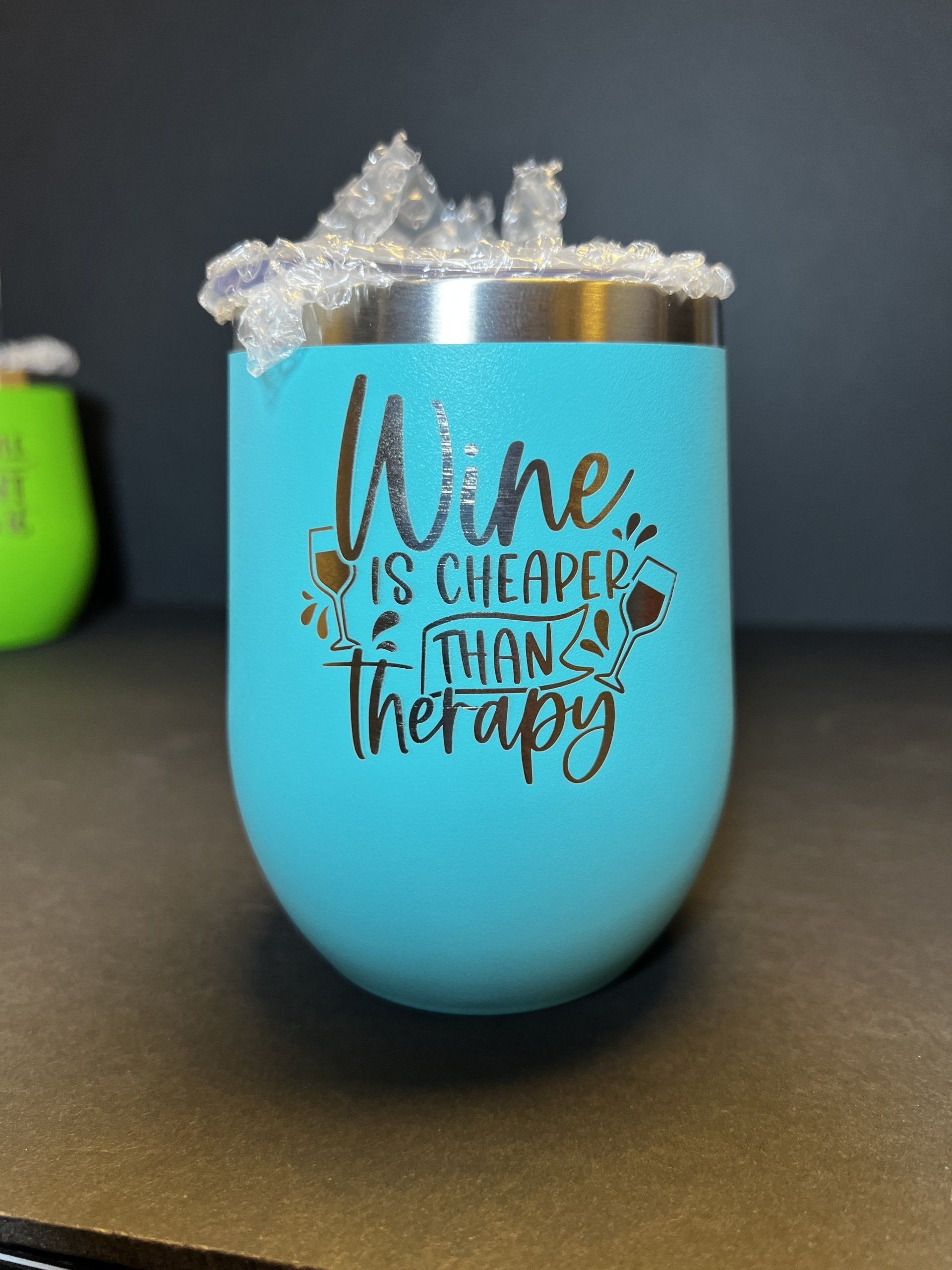 Turquoise wine tumbler with glitter rim and text “Wine is cheaper than therapy” on a dark surface