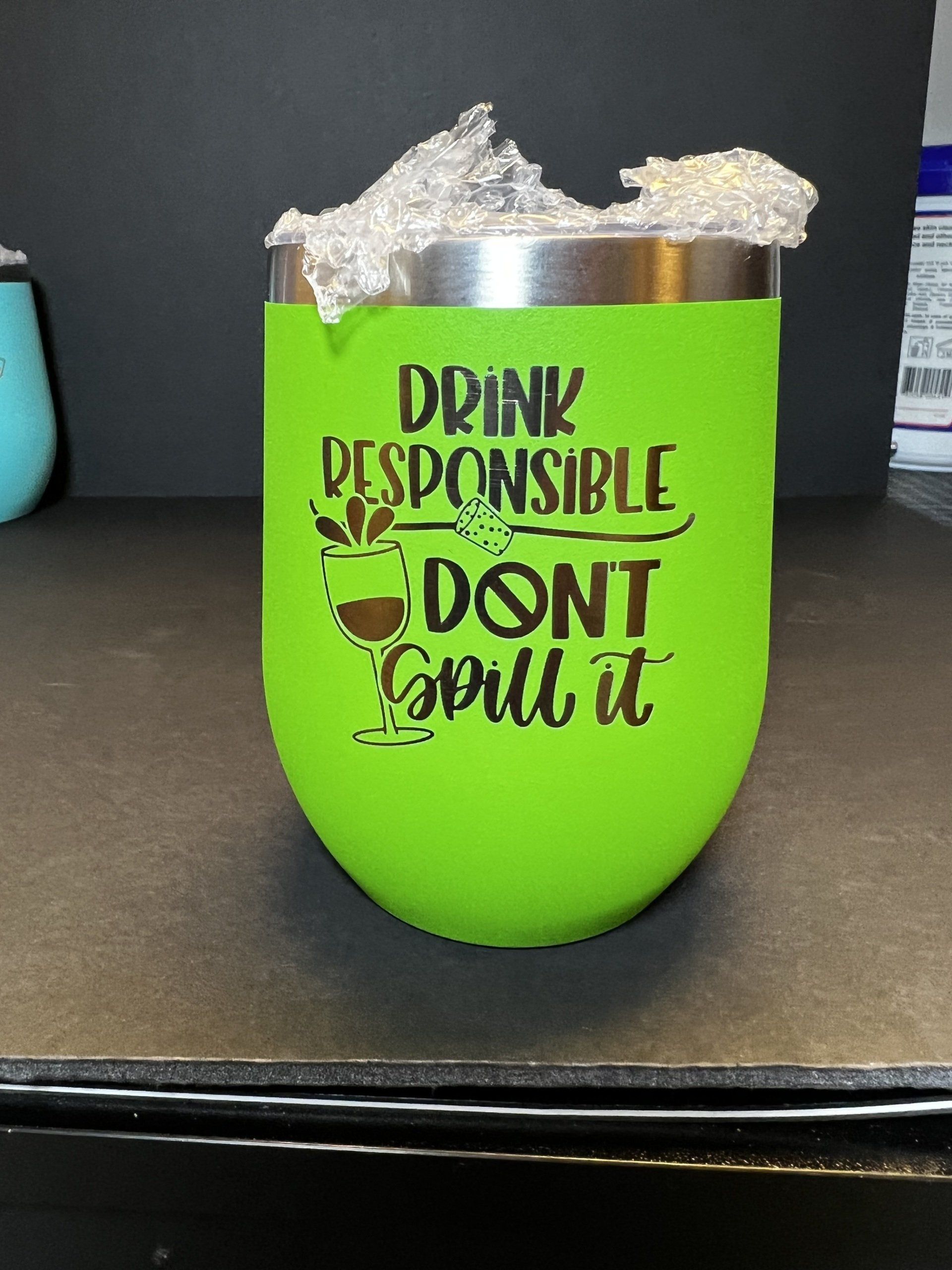 Green koozie with “Drink responsibly, don’t spill it” text holding a silver can on a dark countertop