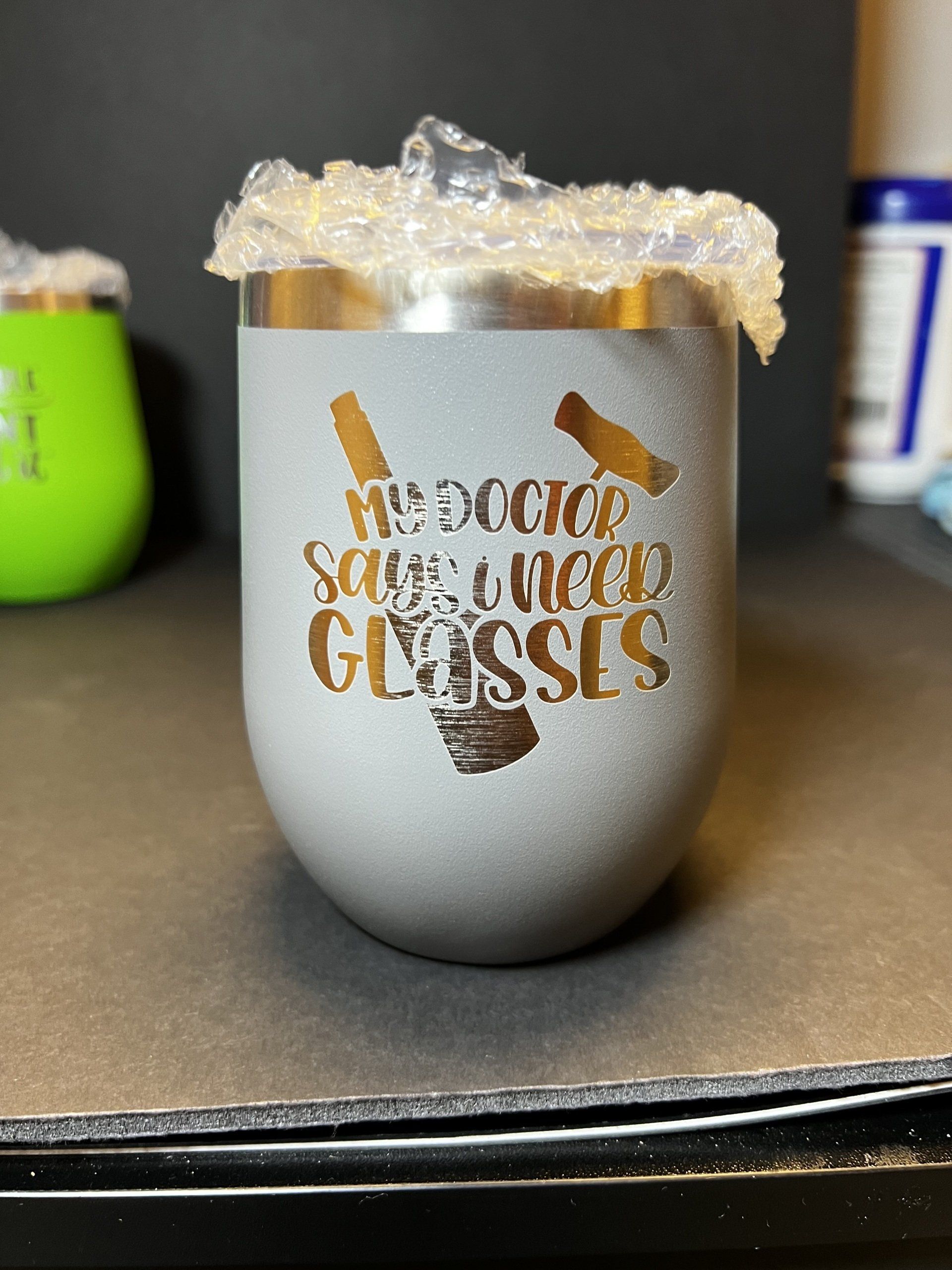 Gray glitter tumbler with gold lettering and a clear plastic wrap on top, sitting on a dark tabletop.