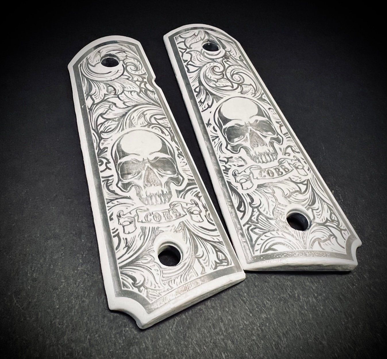 Two engraved silver handgun grip panels with skull and floral designs on a dark surface