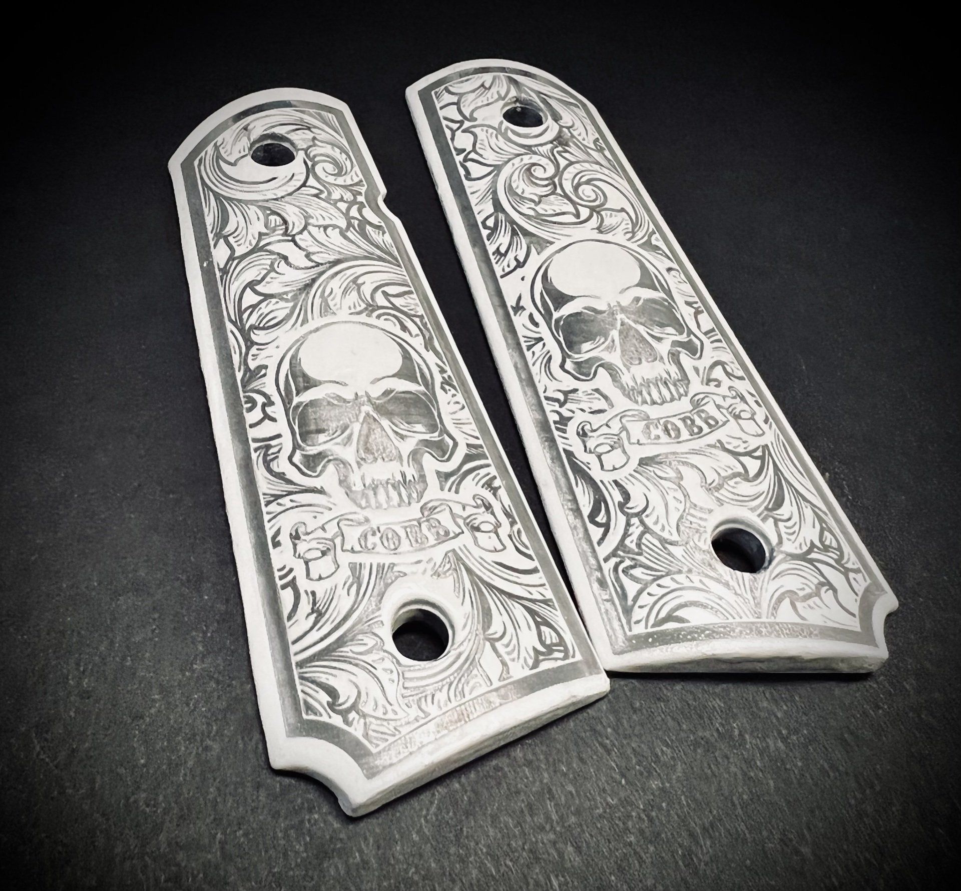 Pair of engraved silver revolver grips with skull and floral designs on a dark background
