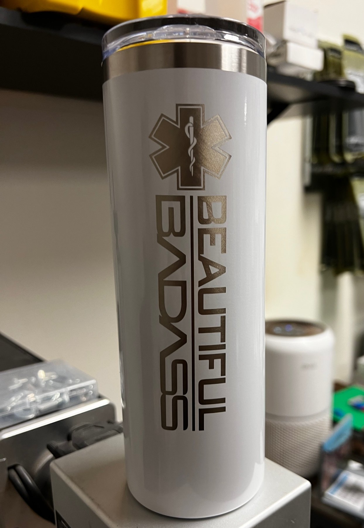 White tumbler with black “Beautiful Badass” text and medical star logo on a desk.