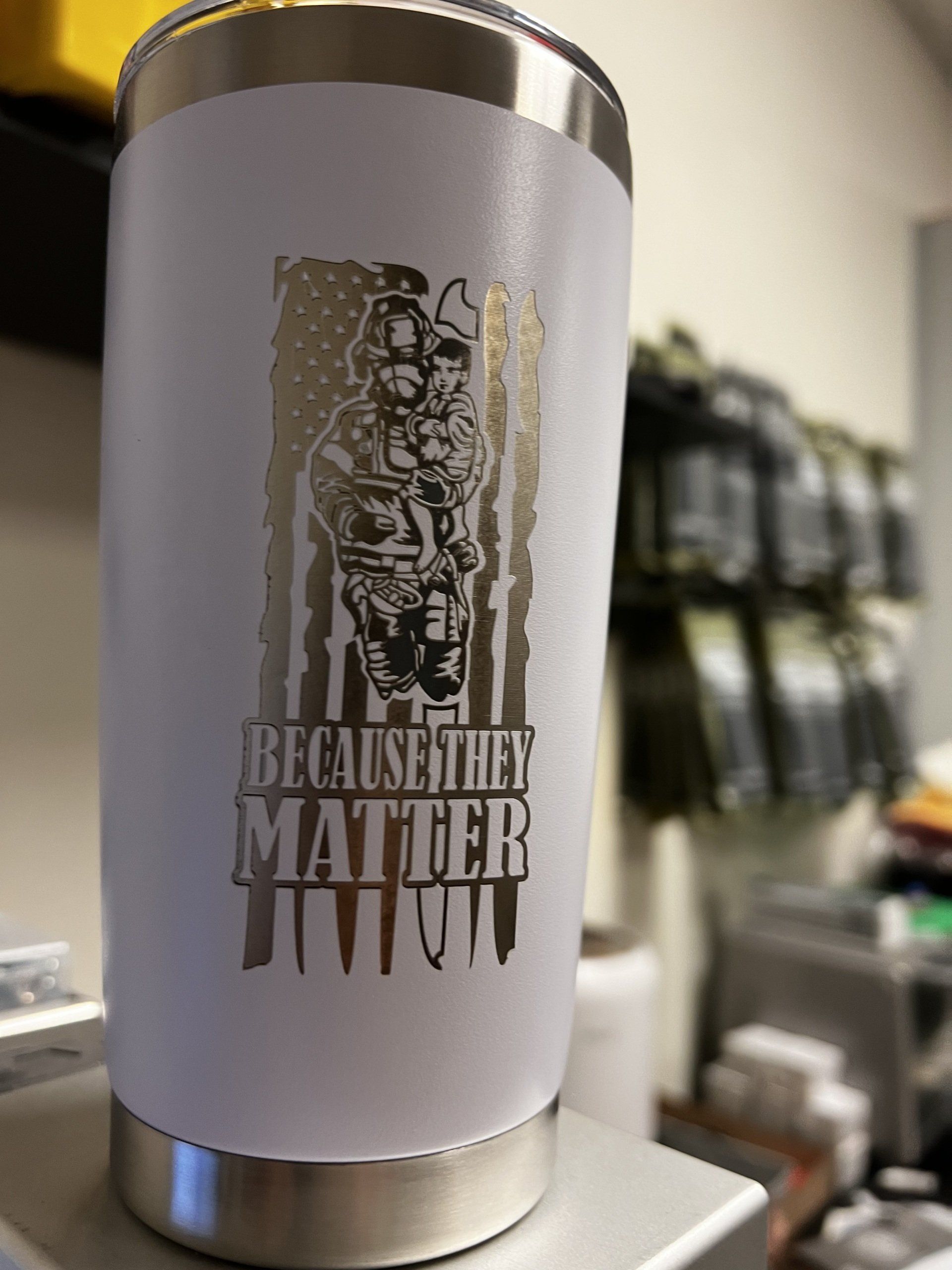 White tumbler with “BECAUSE THE MATTER” text and a graphic on a shop counter, shelves blurred behind it