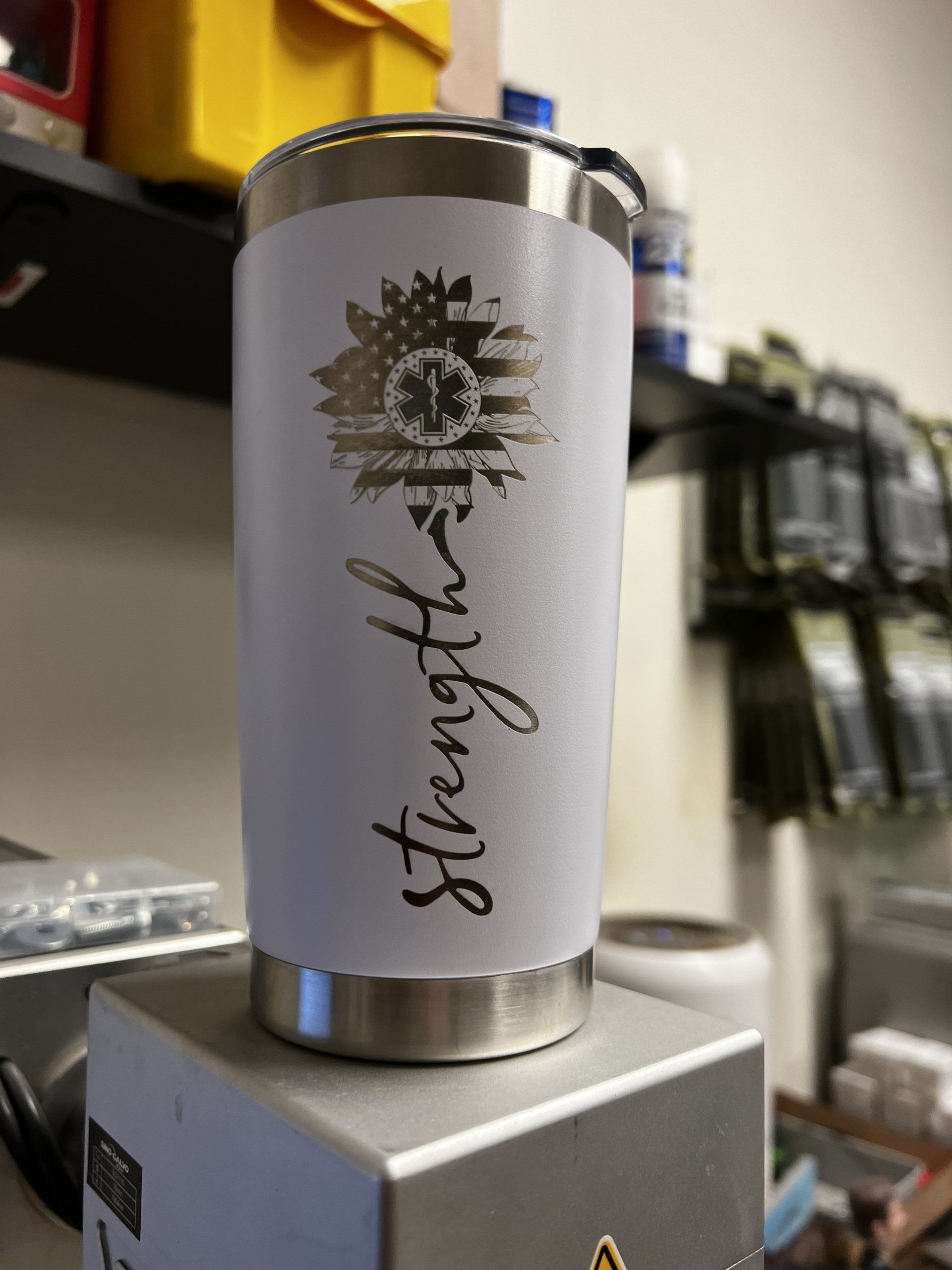 White travel tumbler with black sunburst logo on a gray counter in a shop storage area.