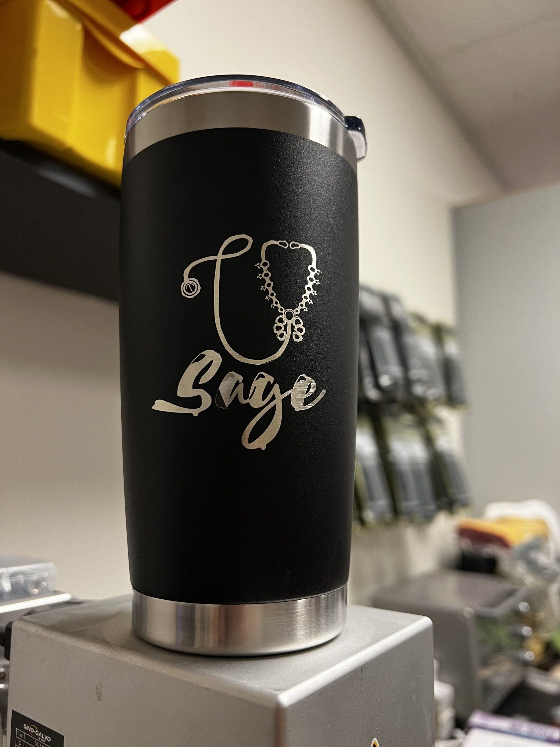 Black insulated tumbler with a stethoscope and “Bage” on a counter in a clinic setting