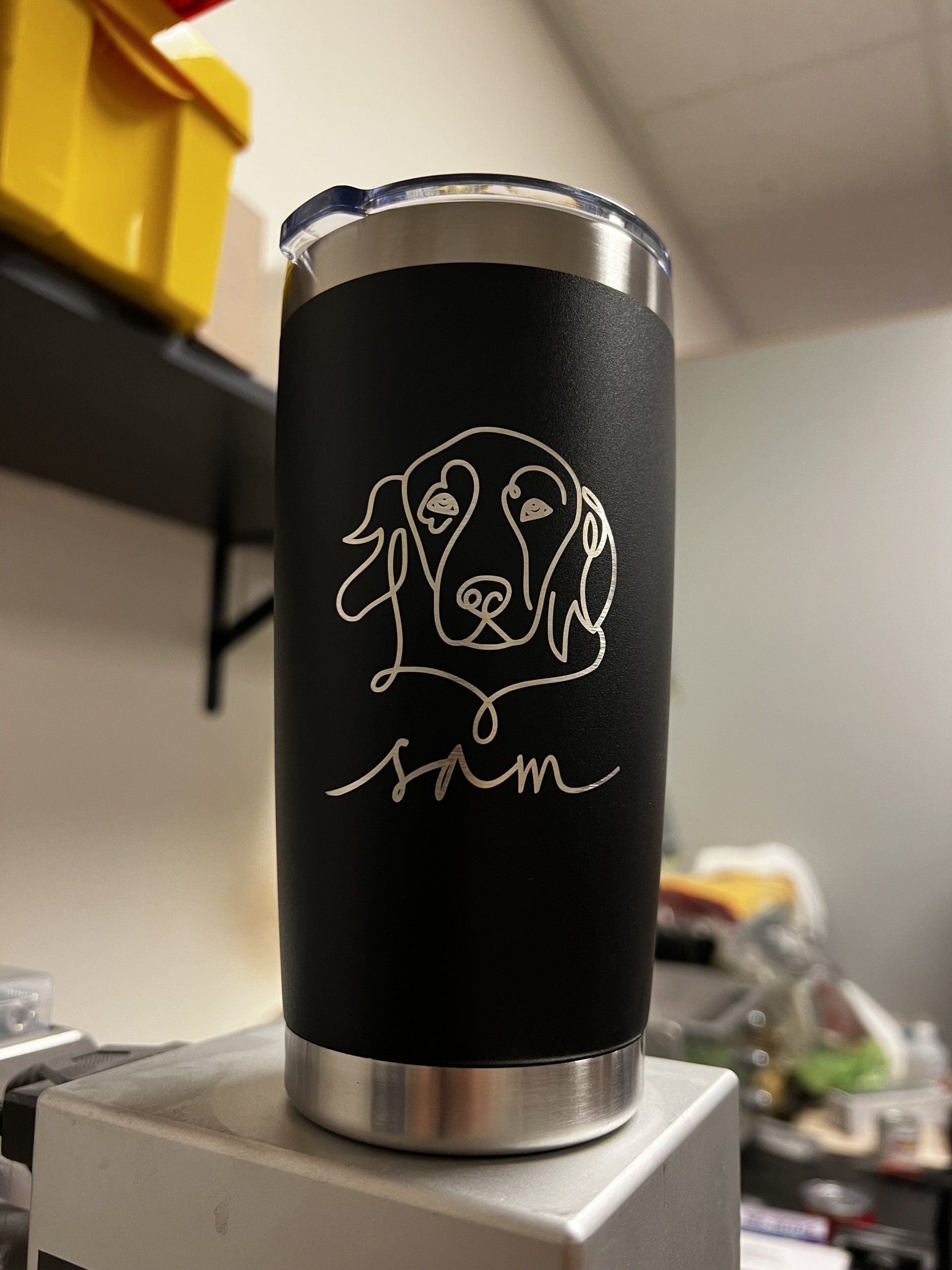 Black insulated tumbler with silver rim and white dog line art, sitting on a box in a room.