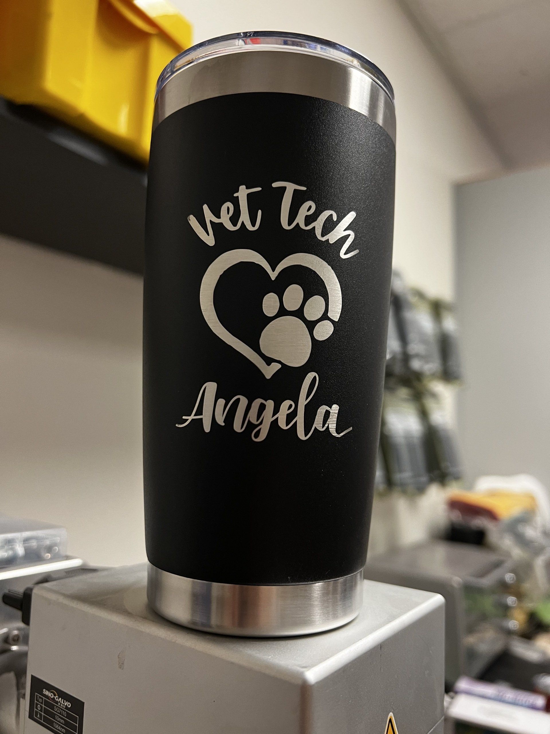 Black tumbler with paw print and “vet tech Angela” on a metal base in a workshop setting