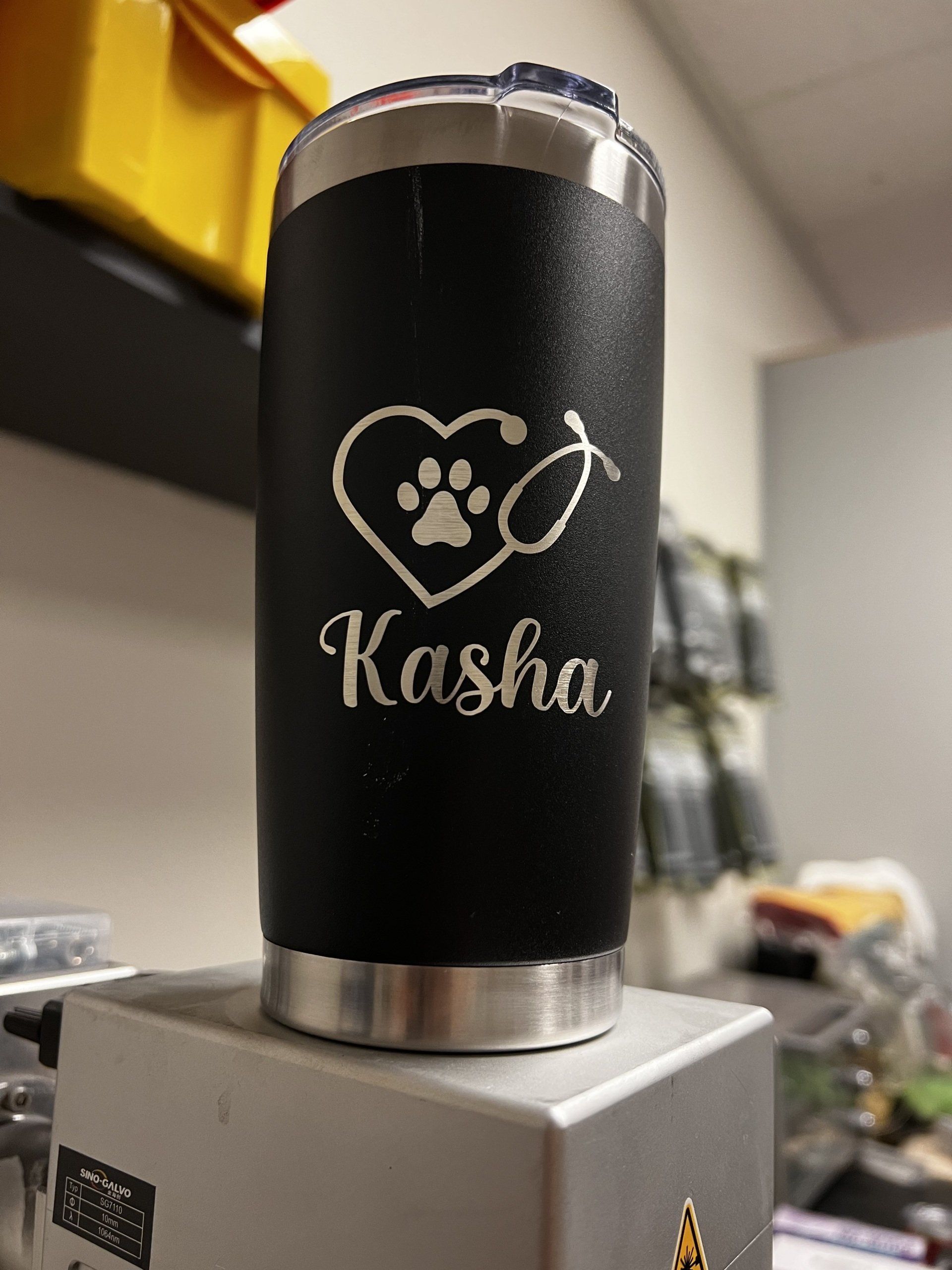 Black tumbler with paw-print heart logo and “Kasha” text sitting on a gray box, indoors.