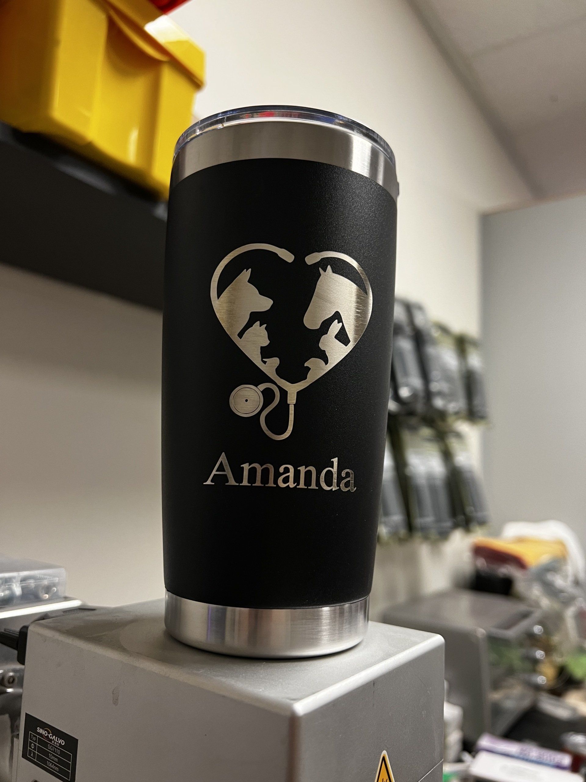 Black stainless tumbler with heart logo and “Amanda” text on a metal surface in a workshop.