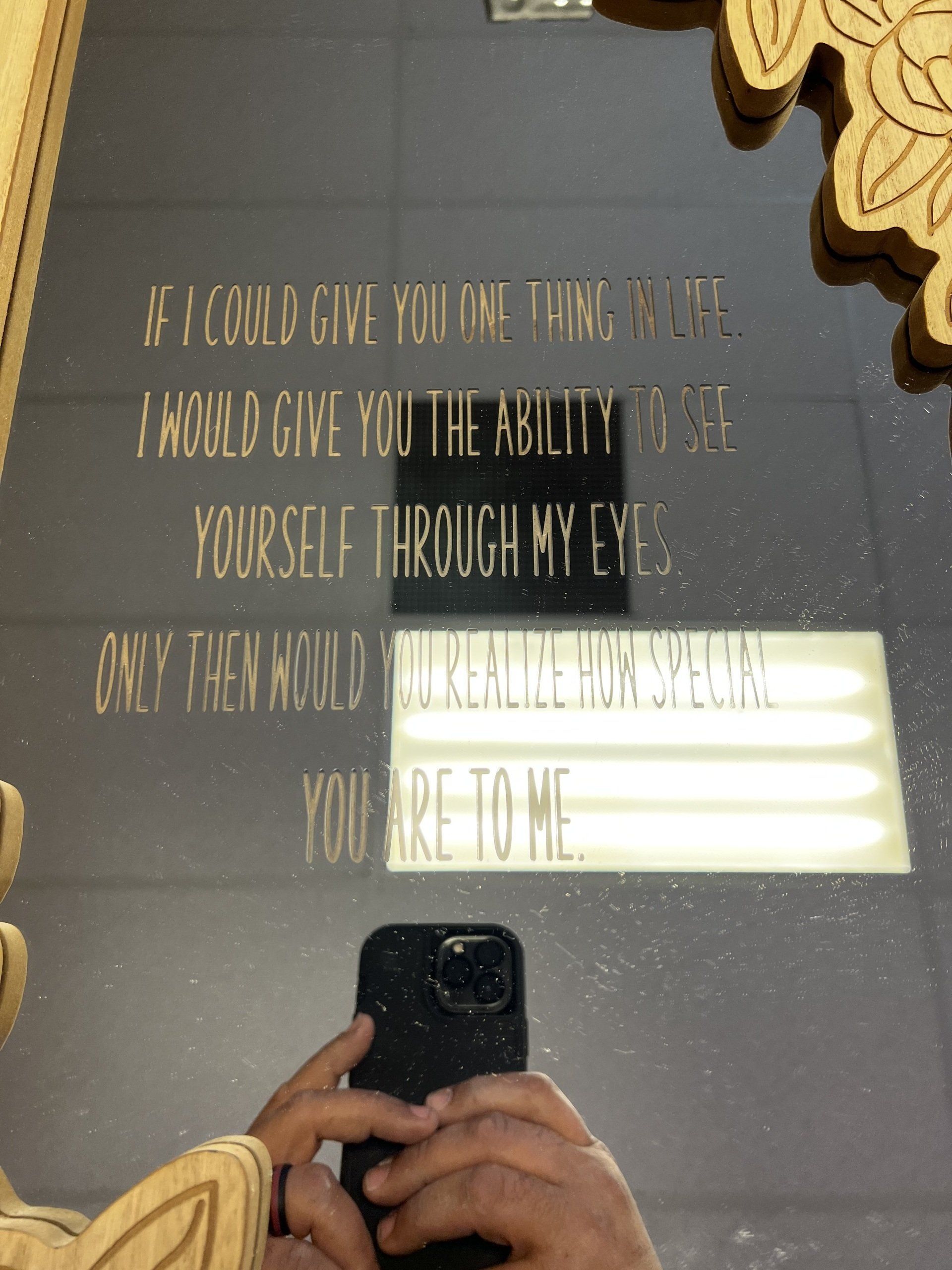 Mirror wall quote in gold lettering, partially obscured by a phone reflection and light glare.