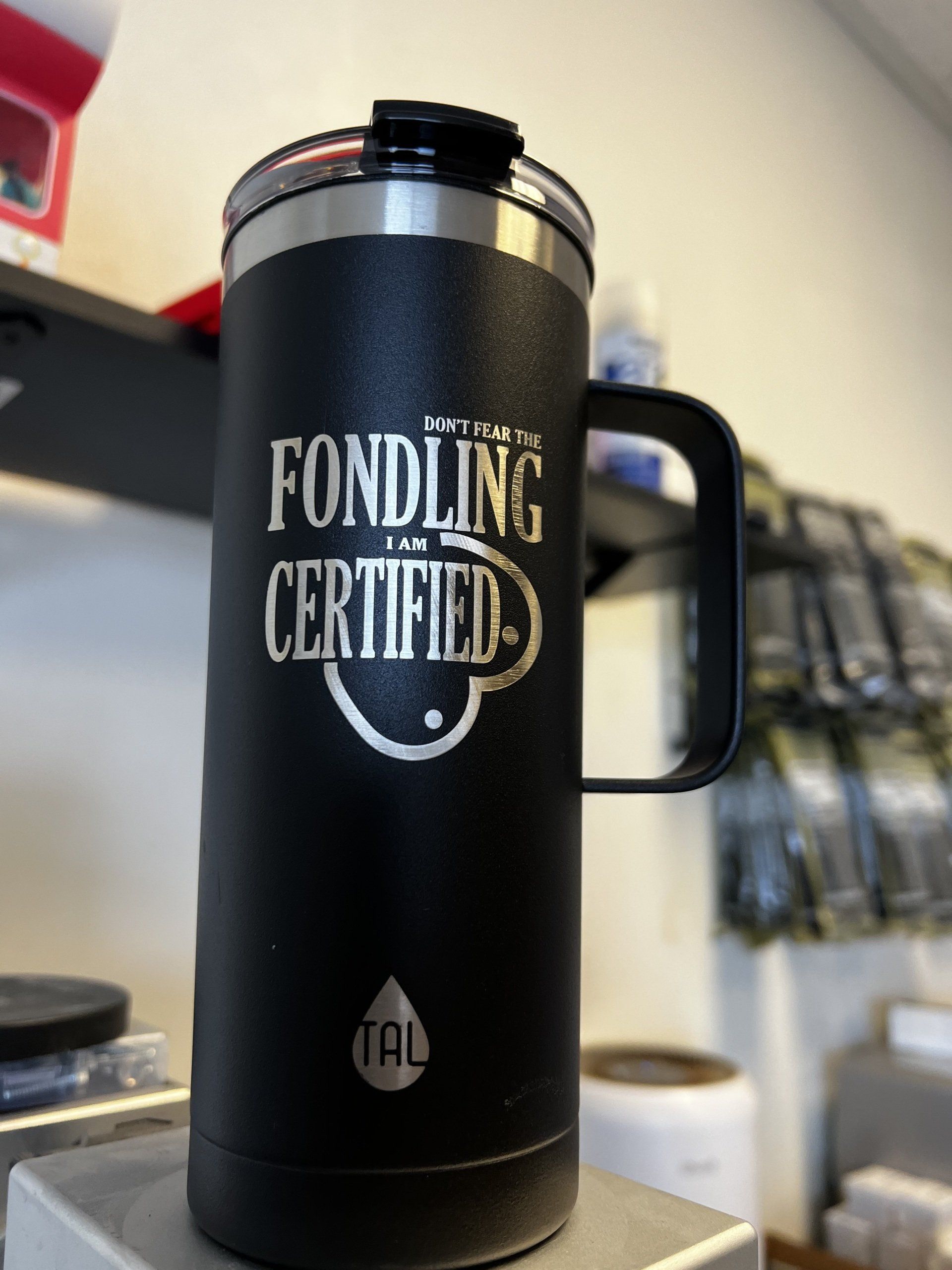 Black insulated tumbler with handle, labeled “Fondling Certified,” on a shelf in a store or café.