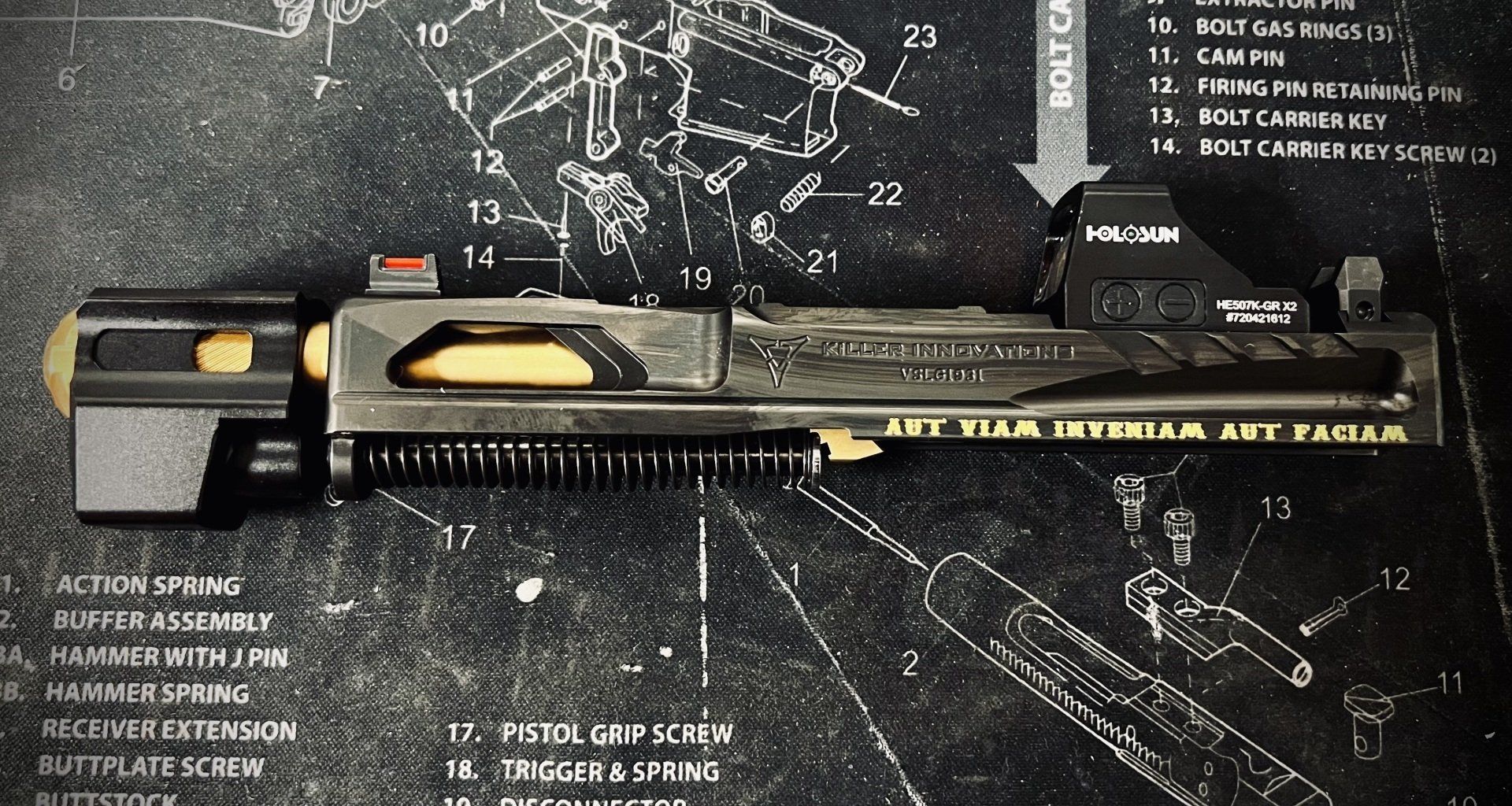 Black-and-gold firearm with scope on a dark technical mat, displayed horizontally.