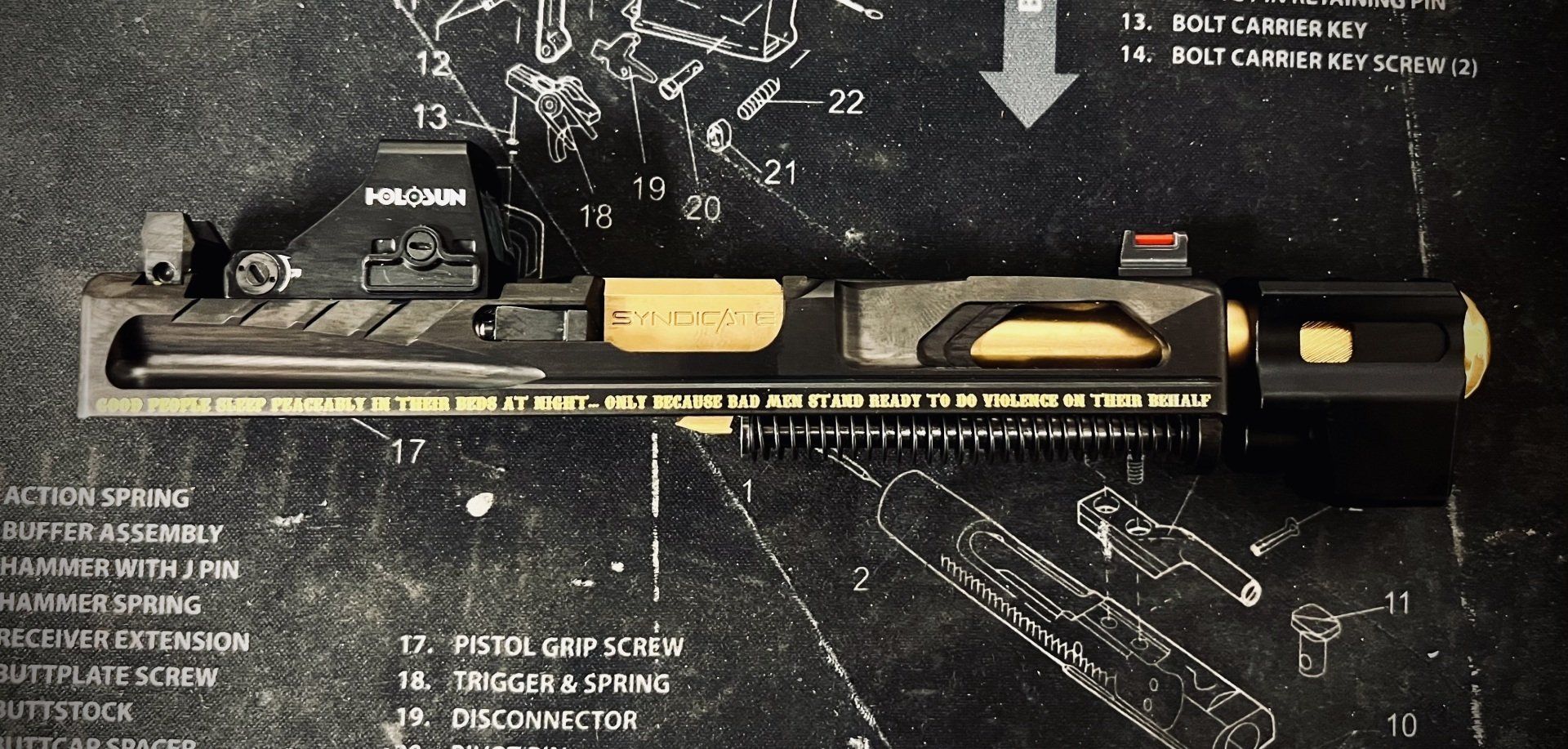 A firearm slide with gold and black elements, mounted with a red dot sight, lies on a gray surface with diagrams.