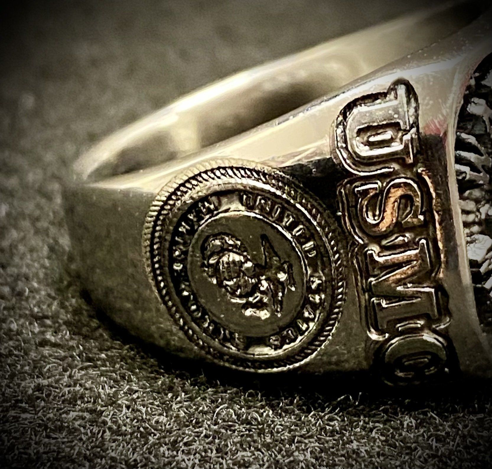Close-up of a silver ring with a circular emblem and raised lettering on a dark textured surface