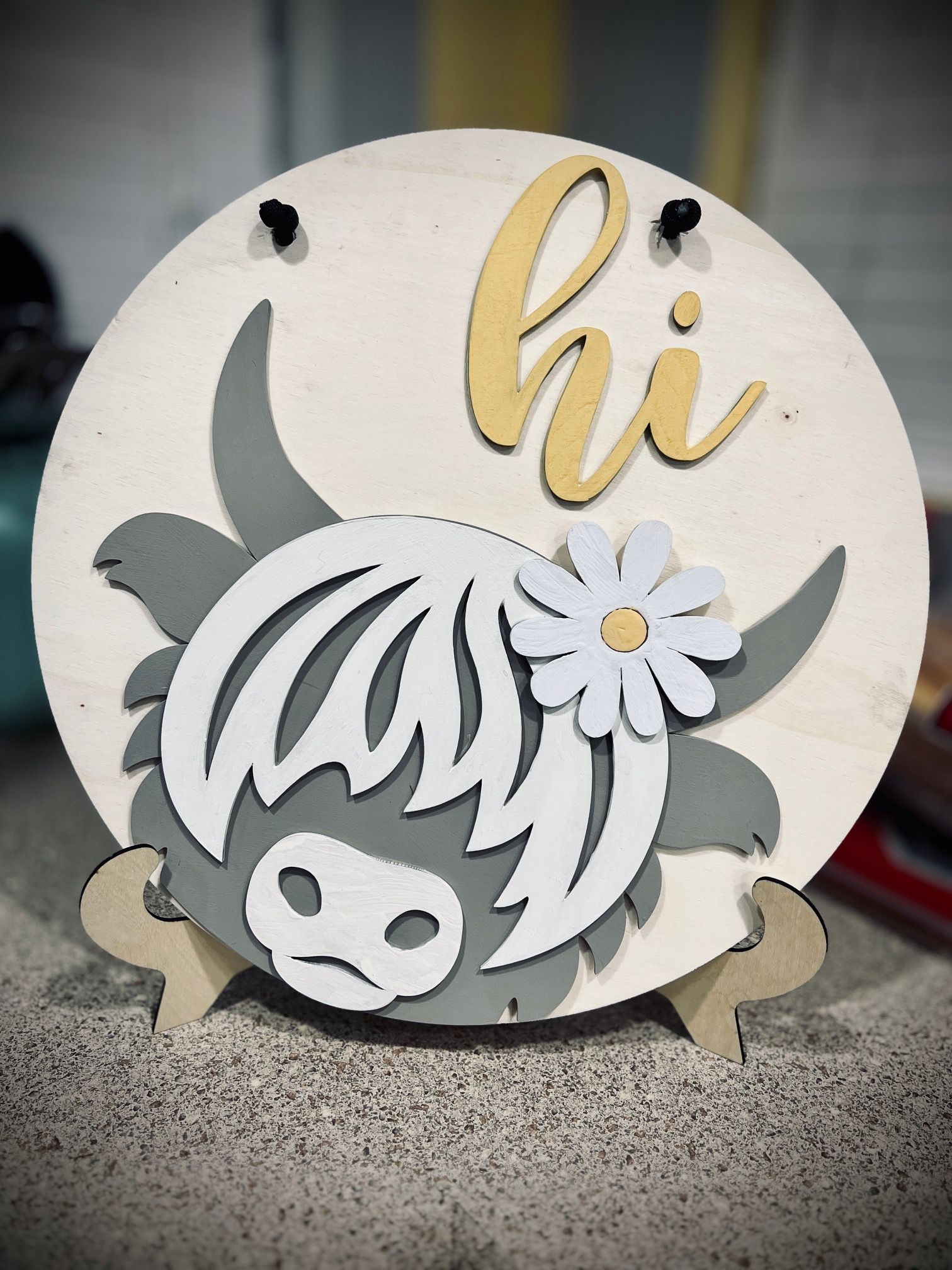Decorative wooden “hi” sign with a gray Highland cow and white flower bow on a round cream plaque.