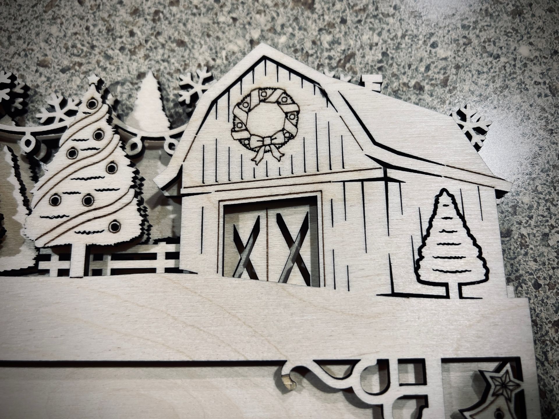Black-and-white paper cutout of a winter barn scene with a wreath, trees, and snowy ground