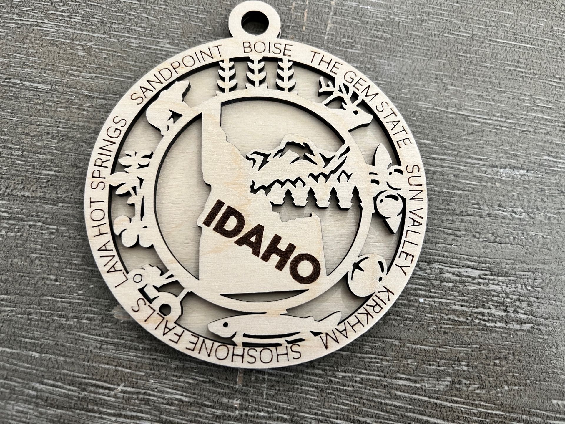 Round Idaho ornament with mountain and forest cutout on a gray fabric surface
