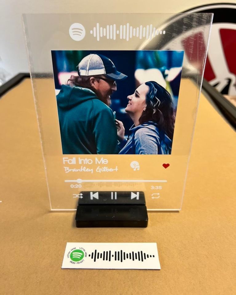 Acrylic photo frame with couple's photo, Spotify code, song title, and play controls. A separate Spotify code sticker rests below.