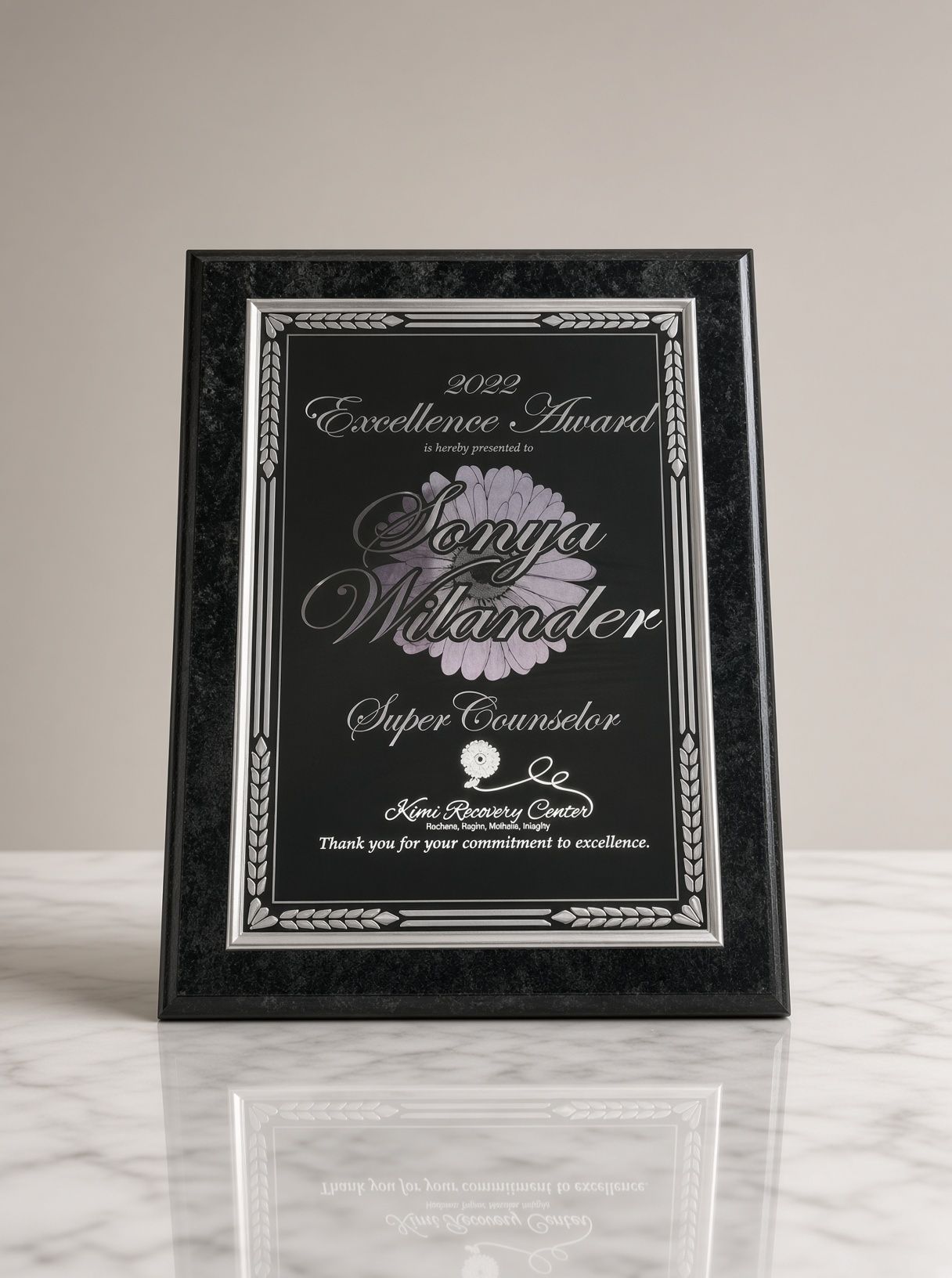 Framed black plaque centered on a Greek laurel wreath design on a light stone background.