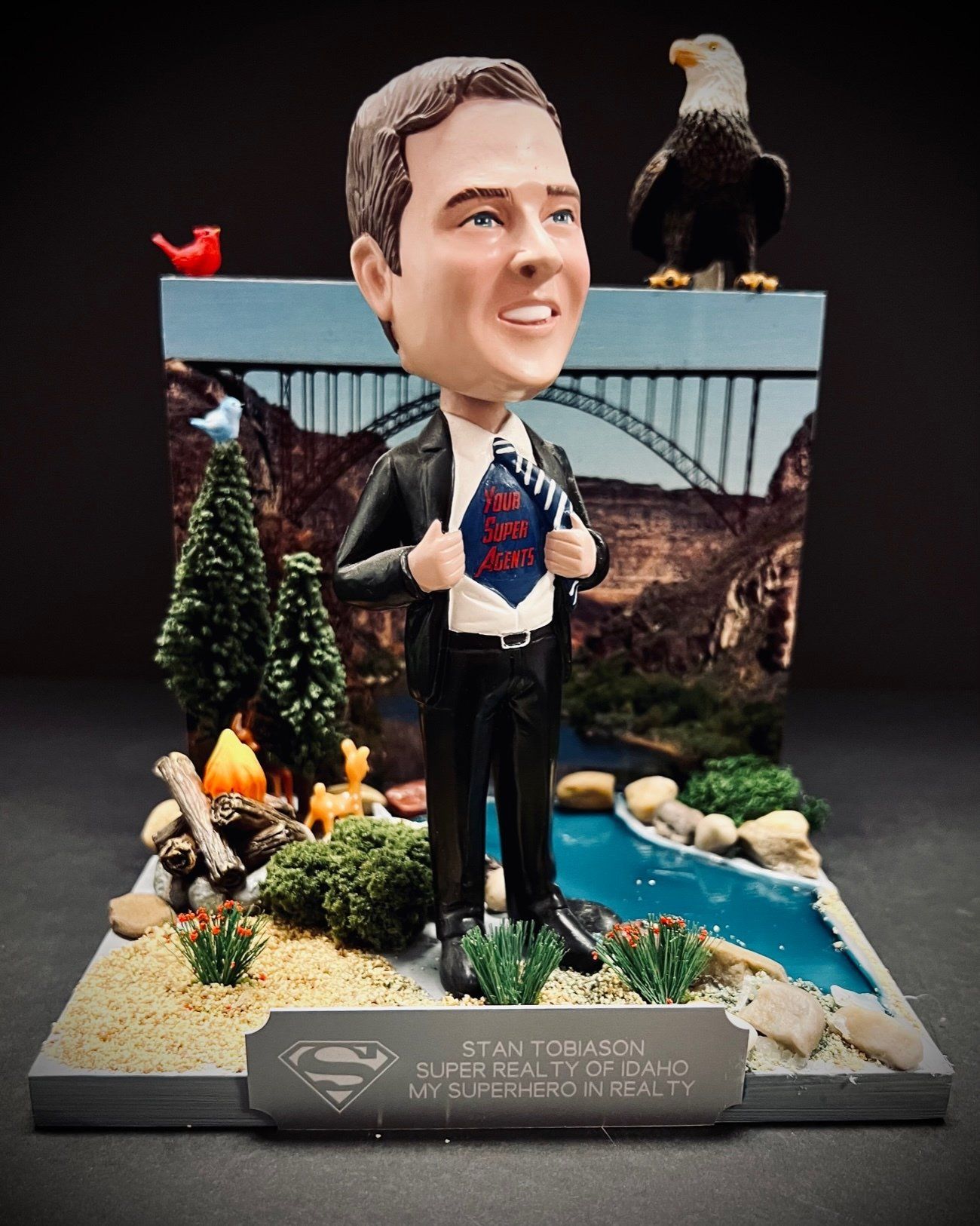 Bobblehead figure in a suit on a scenic diorama with bridge, river, trees, and eagle