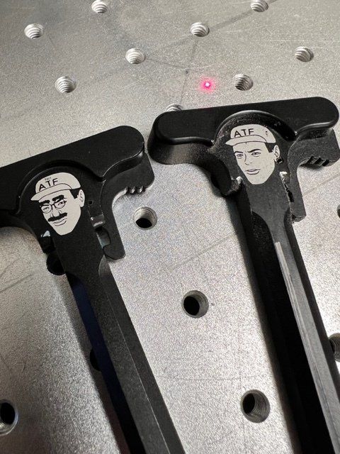 Two black metal gun charging handles with etched portraits of a person wearing a hat. A red laser dot is in the background.