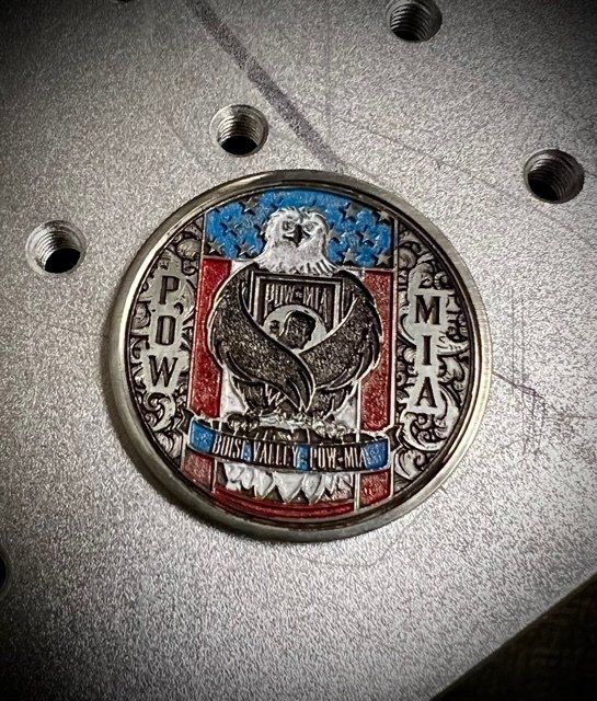 Metal coin with colored POW/MIA symbol of an eagle with shield, on metal surface.