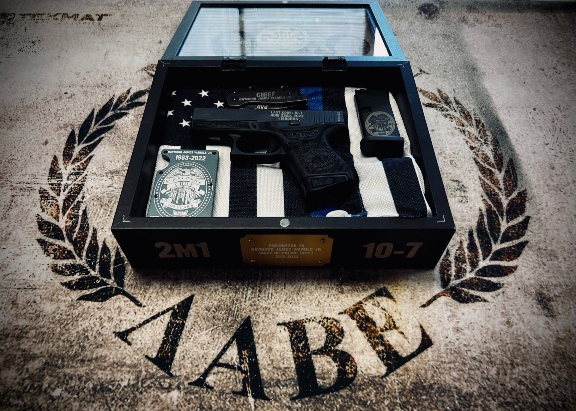 Gun and police items in a memorial shadow box: pistol, watch, badge, flag, and a laurel wreath.