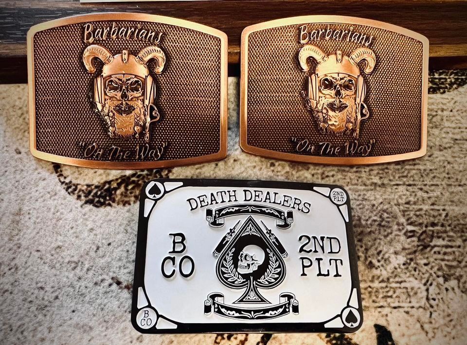 Three belt buckles: two copper-toned with a bear helmet design, and one silver with a spade and text that reads 