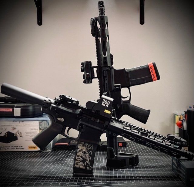 Two black rifles with optics on a table, one with a suppressor and the other with a red magazine.