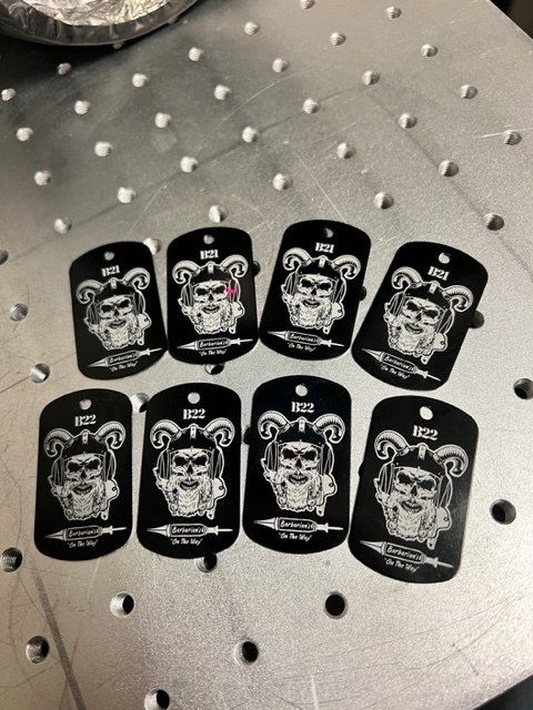 Eight black metal dog tags with a skull graphic and text, laid out on a perforated silver surface.