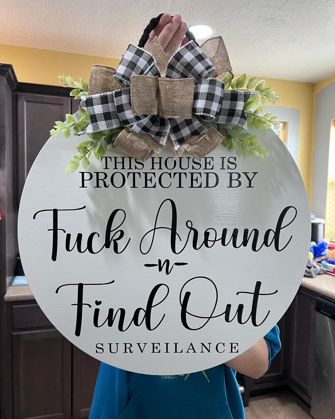 Round sign with plaid bow reads “This house is protected by Tuck Around and Find Out Surveillance.”