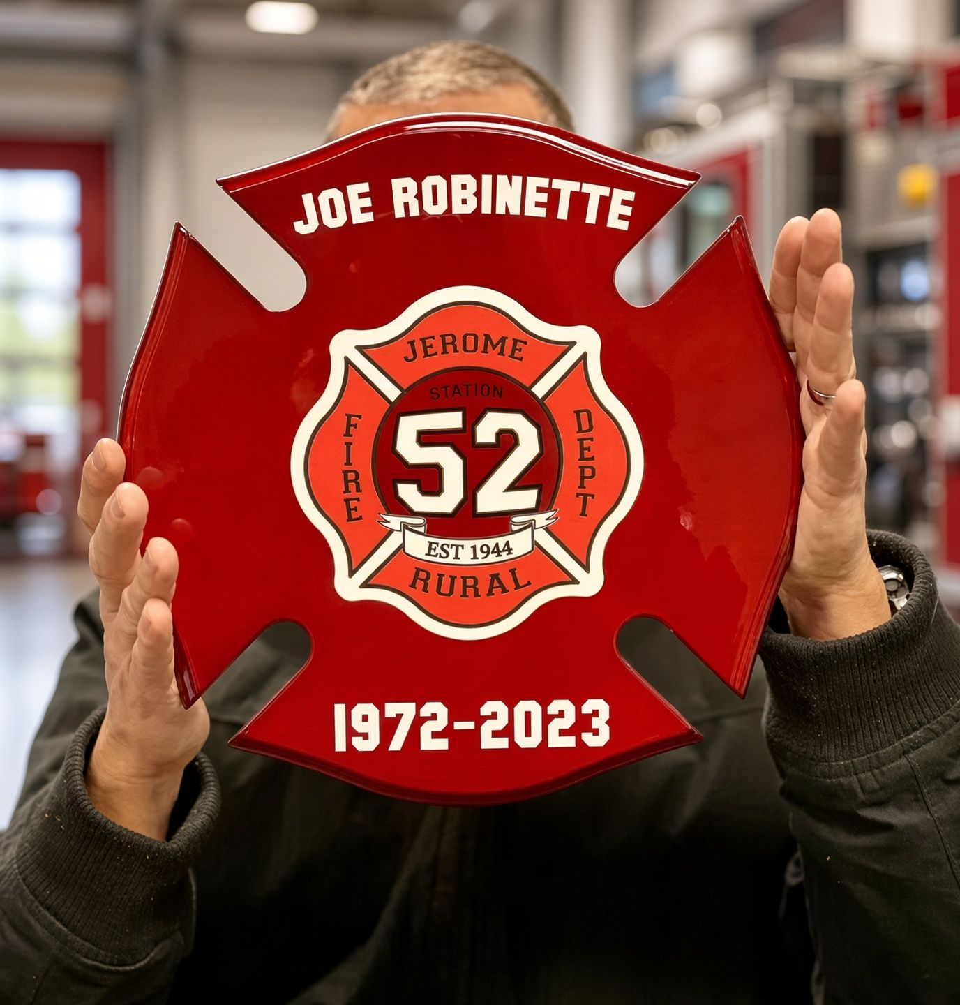 Red firefighter memorial plaque with “Joe Robinette,” “Jerome Hural,” “52,” and “1972–2023” held by two hands