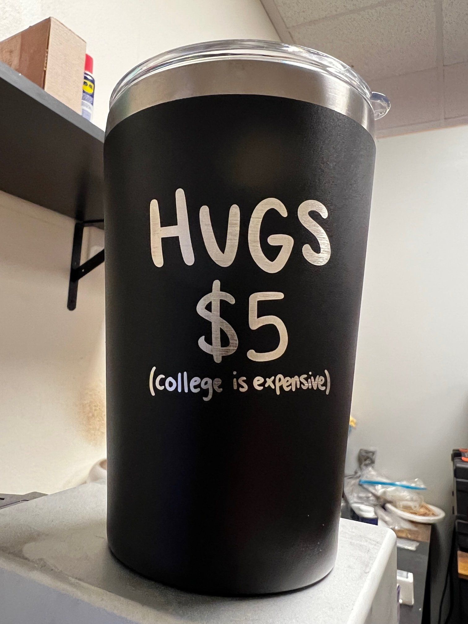 Black donation bin labeled “HUGS $5 (College is Expensive)” in an indoor room