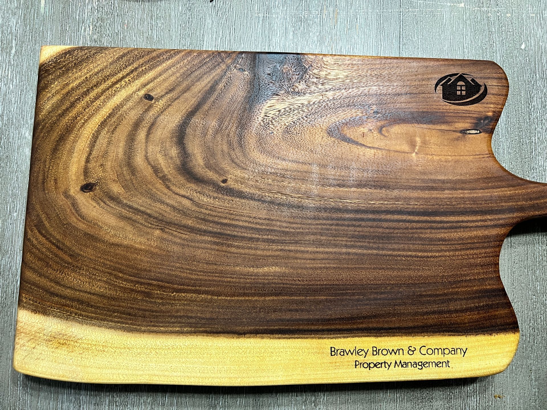 Wooden cutting board with dark grain and a light base, sitting on a textured surface.
