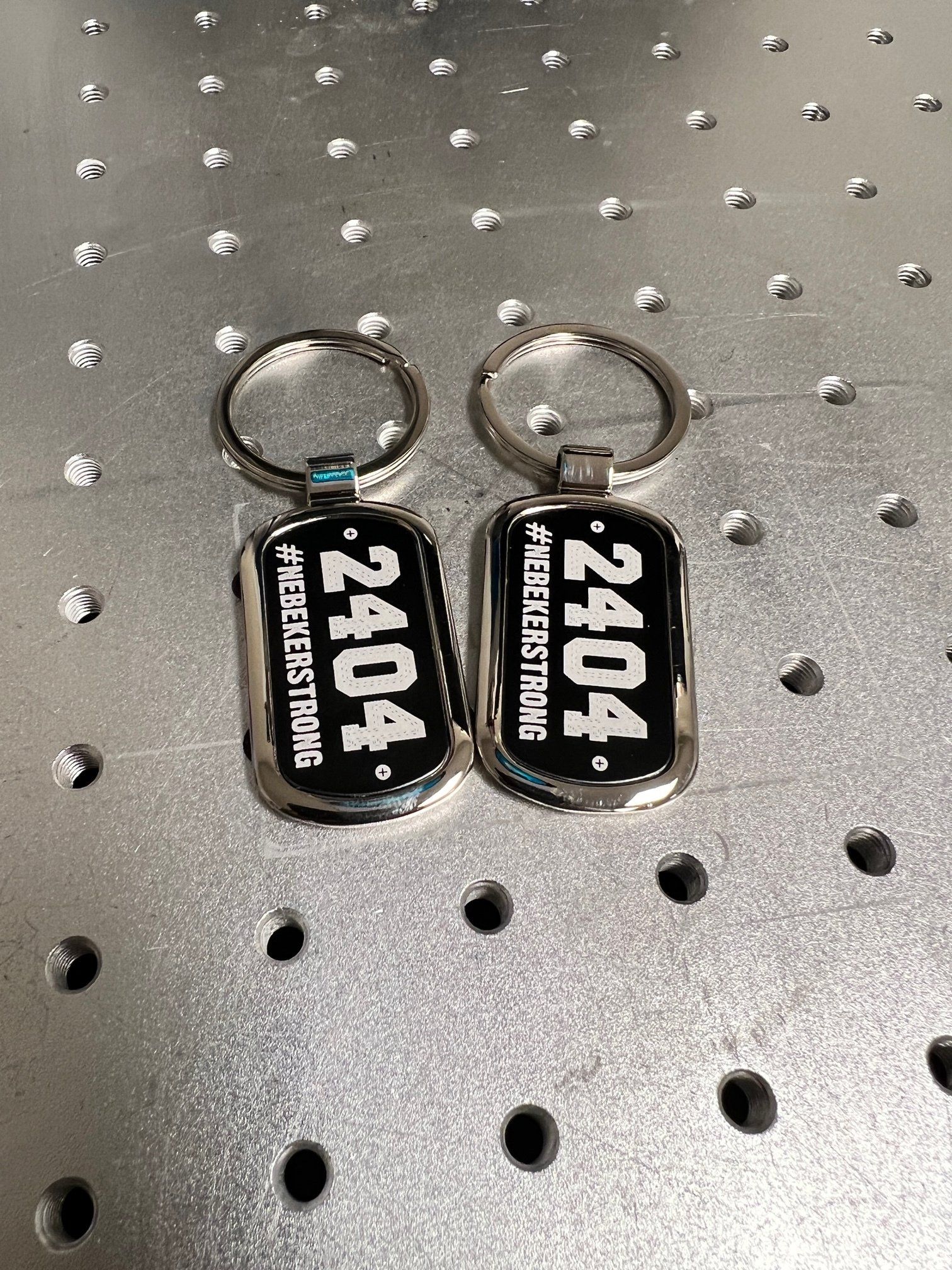 Two engraved keychains on a metal surface, both with black backgrounds and silver trim. Each displays 