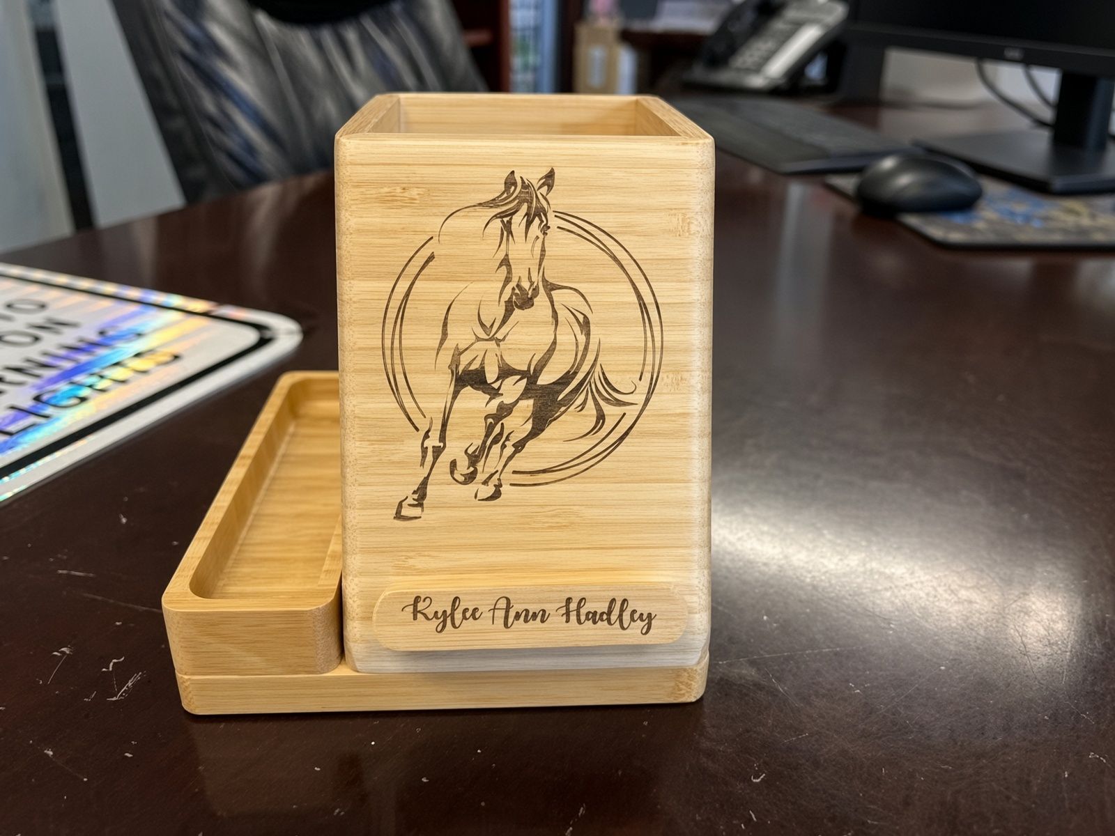 Wooden phone stand engraved with a horse sketch, on a desk with a tray and papers nearby.