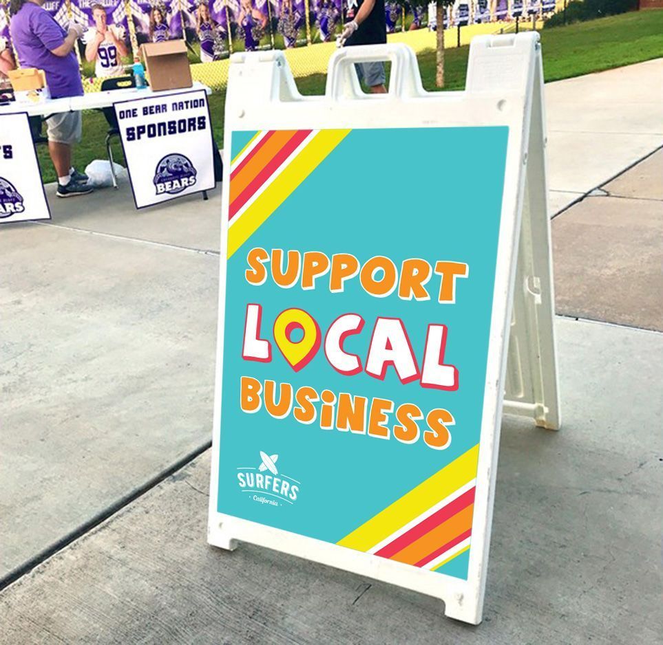 A teal sidewalk sign reads “Support Local Business” in colorful letters near a sports venue.
