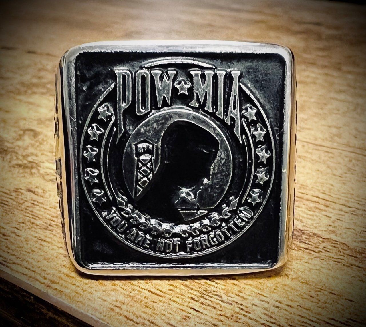 Black square metal badge with embossed circular logo on a wooden surface, lit warmly.