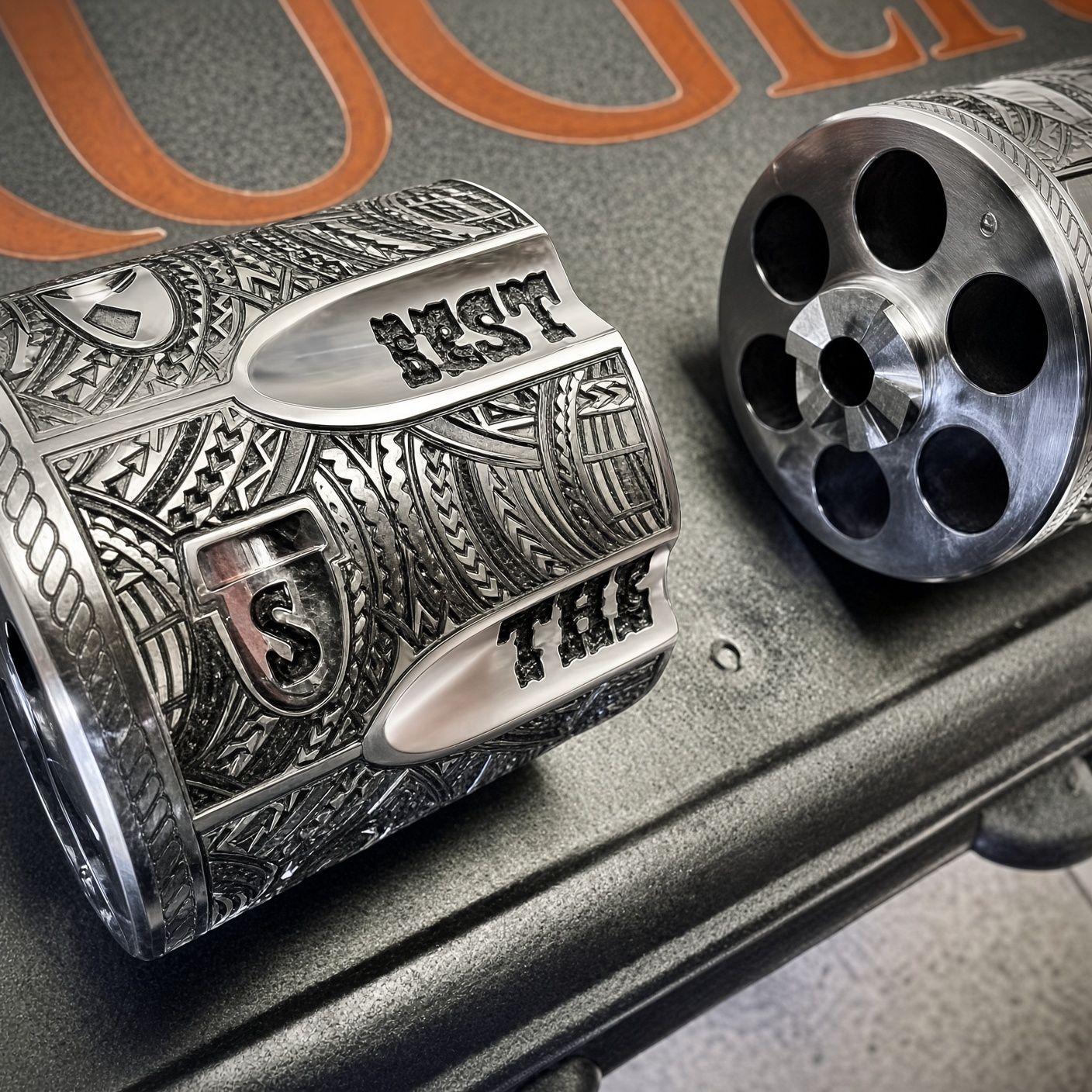 Close-up of a engraved silver revolver cylinder with ornate dollar-bill design on a metallic surface