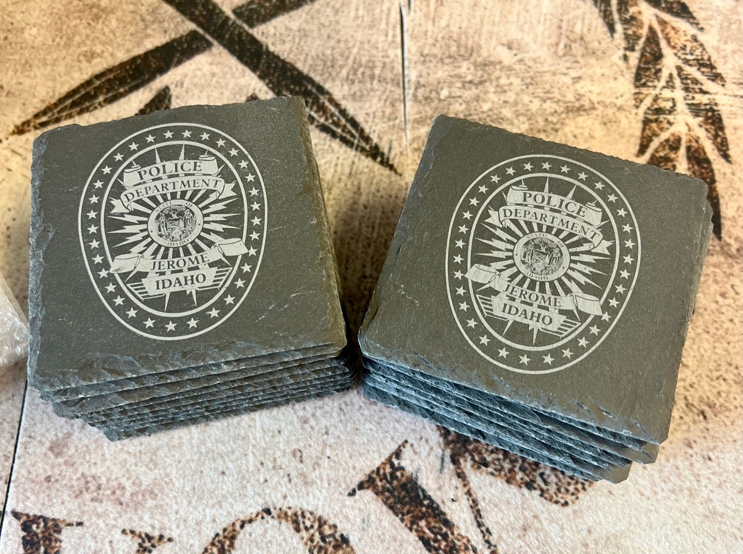 Two stacks of dark square coasters with white circular designs on a patterned cloth surface