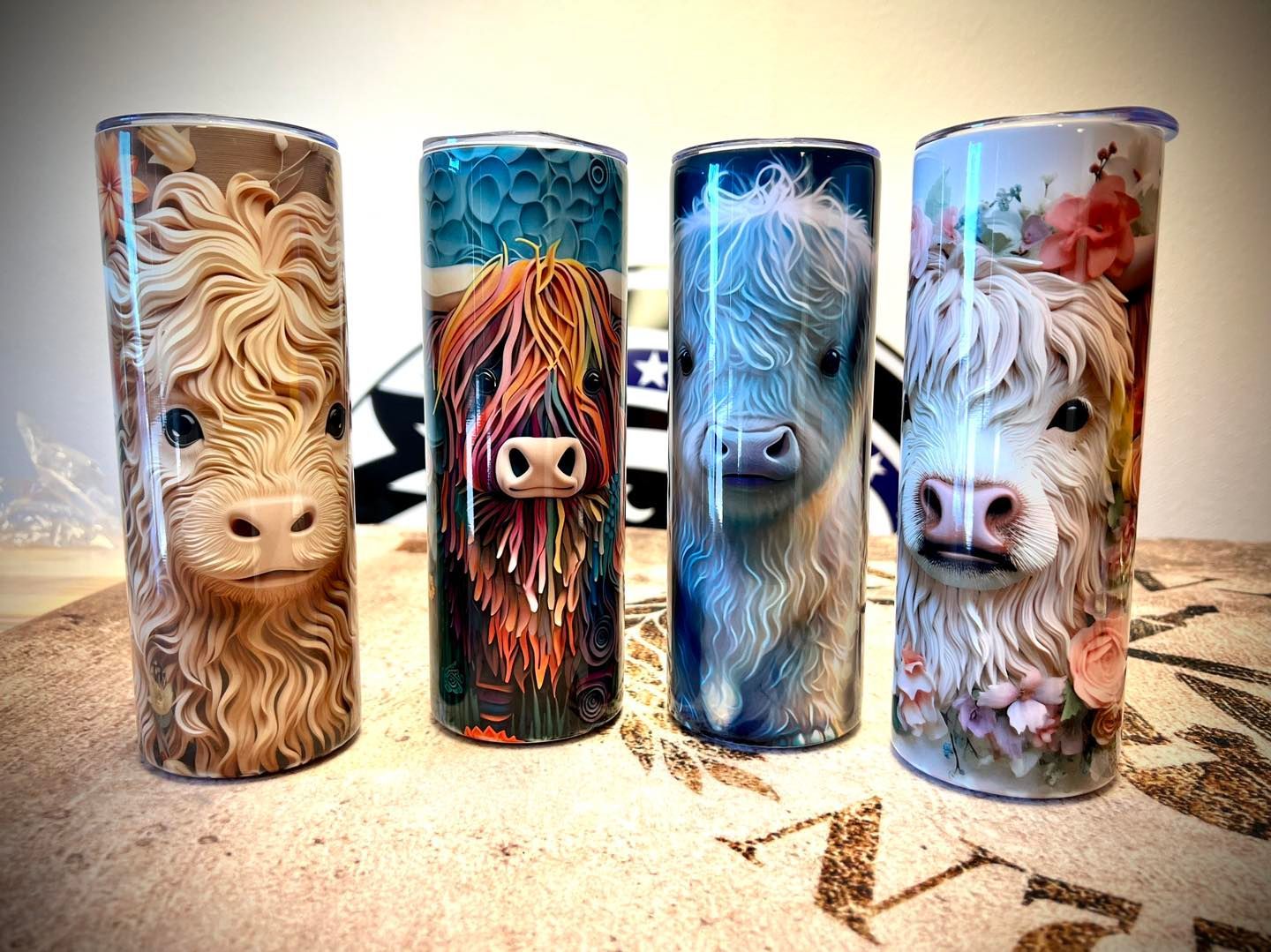 Four cylindrical tumblers with highland cow designs. Various color schemes: brown, orange, blue, floral.