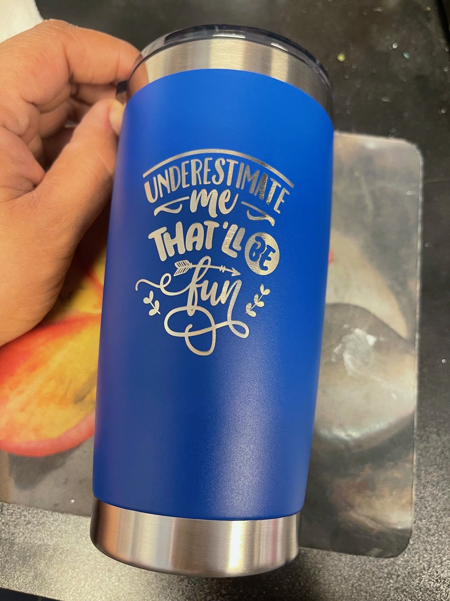 Blue insulated tumbler with white “Underestimate me, that’ll be fun” lettering held in a hand