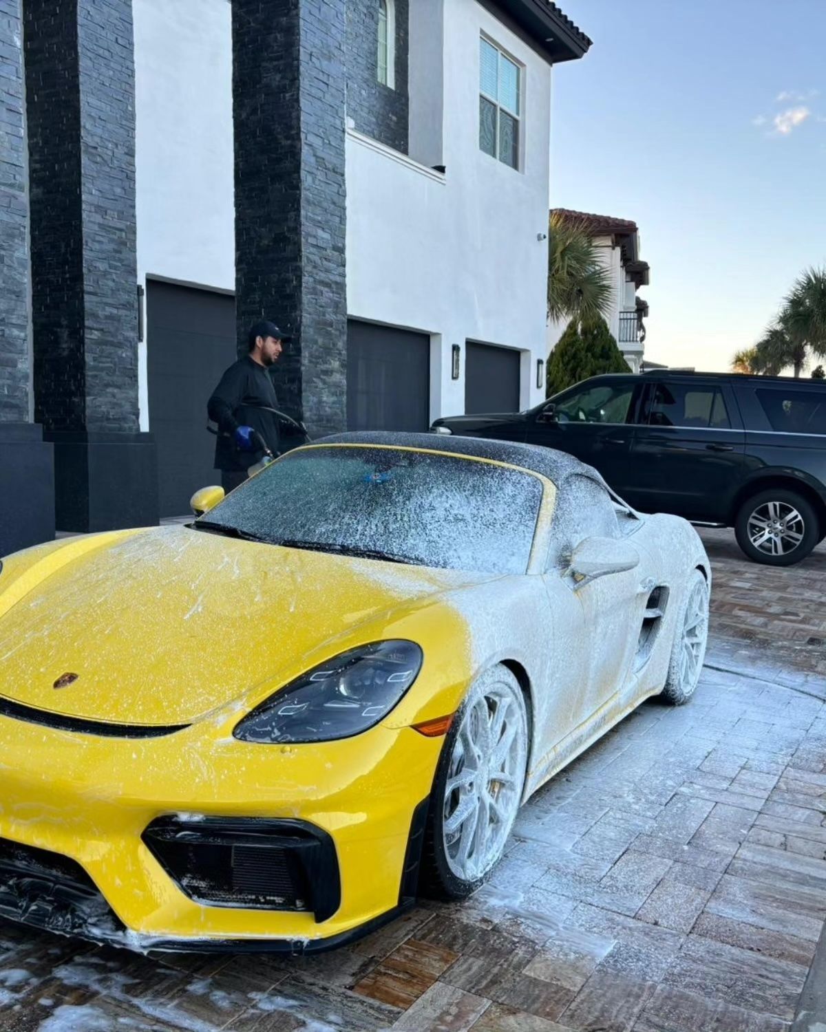 A yellow and white Porsche being washed by a person in front of a modern house.