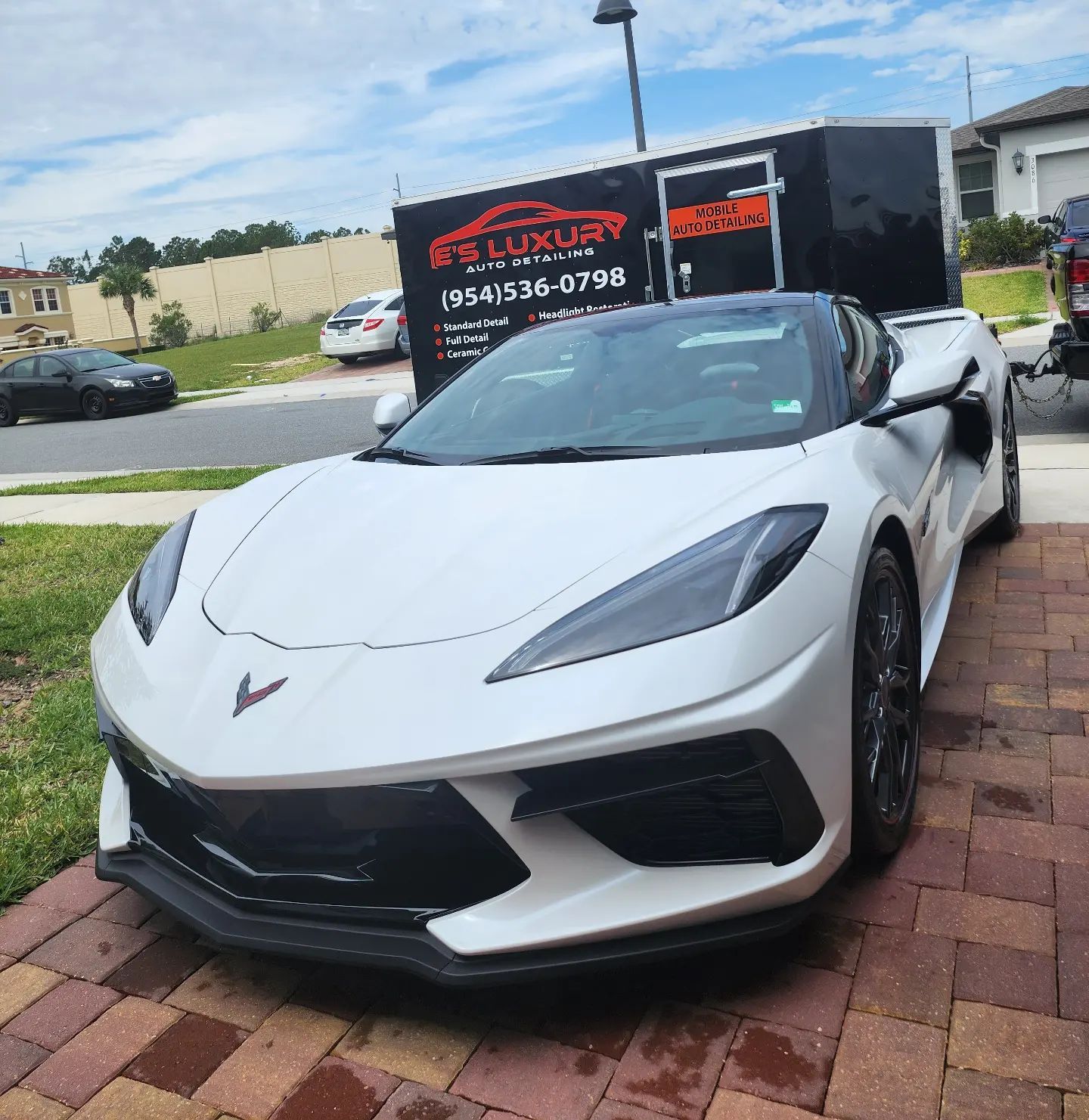 White sports car parked on brick, in front of black trailer; daytime.
